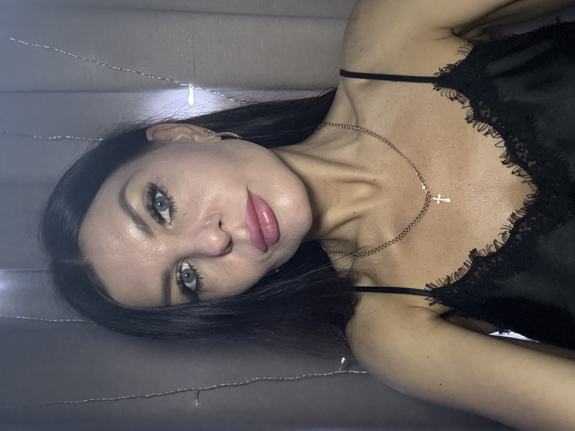 A woman with dark hair is lying on a gray surface. She is wearing a black top with lace trim and a gold necklace with a cross pendant. She has light-colored eyes and pink lipstick. There are small string lights in the background. There is no evidence to suggest the location is Chaikovsky, Russia.