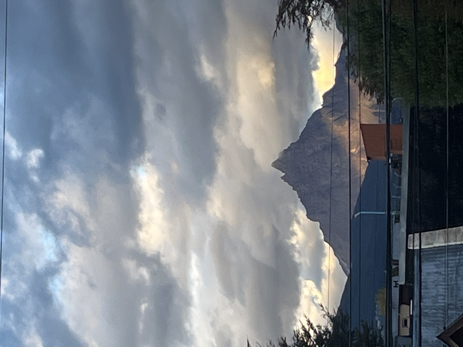 The image displays a static outdoor scene in Ushuaia, Argentina, centered on a prominent, dark grey mountain peak identifiable as Monte Olivia. The upper two-thirds of the frame are dominated by a partly cloudy sky, featuring dense, dark grey clouds intermixed with illuminated patches of pale yellow and bright white where sunlight penetrates. A single, thin, dark utility wire crosses horizontally at the extreme top. Monte Olivia rises sharply in the midground, its rocky, jagged form exhibiting areas of shadow and a distinct shaft of yellowish sunlight illuminating its lower right slope. Below the mountain, the foreground reveals urban architectural elements: a dark-colored building roof with a lighter ridge line, a small, rust-orange rectangular structure to its right, and a partial dark blue structure on the left. Multiple dark-colored utility wires extend horizontally across the lower third of the image, positioned above these foreground structures. Patches of dark green tree foliage are visible at the far left and right edges of the frame. No individuals or active subjects are present.