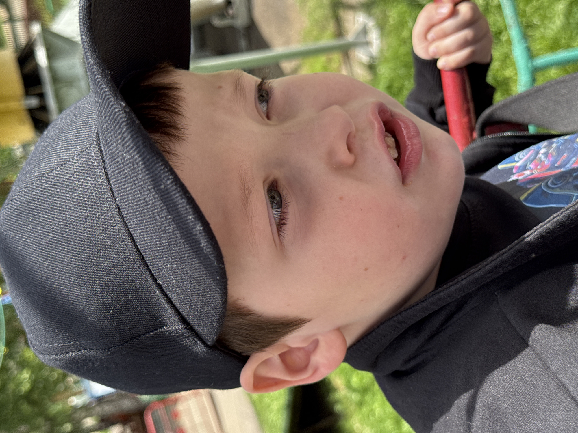 A close-up, outdoor, eye-level, medium shot shows the right side of a young boy's face as he looks to his right, with his mouth slightly open. He wears a black baseball cap and a black jacket over a graphic t-shirt. His skin is fair with some freckles. The background is blurred and appears to be a park or playground, with green grass and some structures that suggest the city of Chaikovsky, Russia. The boy's right hand is visible, gripping a red bar.