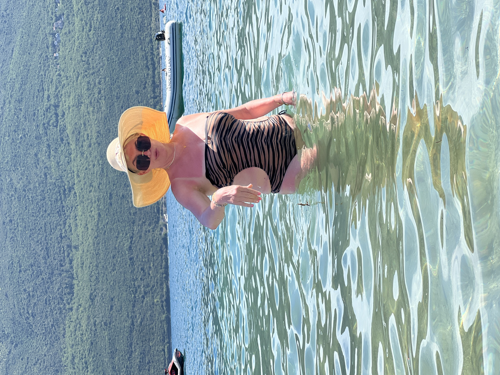 A woman stands in the clear, shallow water of the Bay of Kotor, near Baošići, Montenegro. She is wearing a tan and black tiger-striped one-piece swimsuit, a wide-brimmed straw hat, and sunglasses. Her left arm is extended slightly forward, with her hand open. In the background, a green, tree-covered mountainside rises from the water, with a few houses visible near the top. A white inflatable boat with "TENDER TO LANA" printed on its side is moored in the water, and a jet ski is visible to the left. The water is calm with ripples reflecting the sunlight.