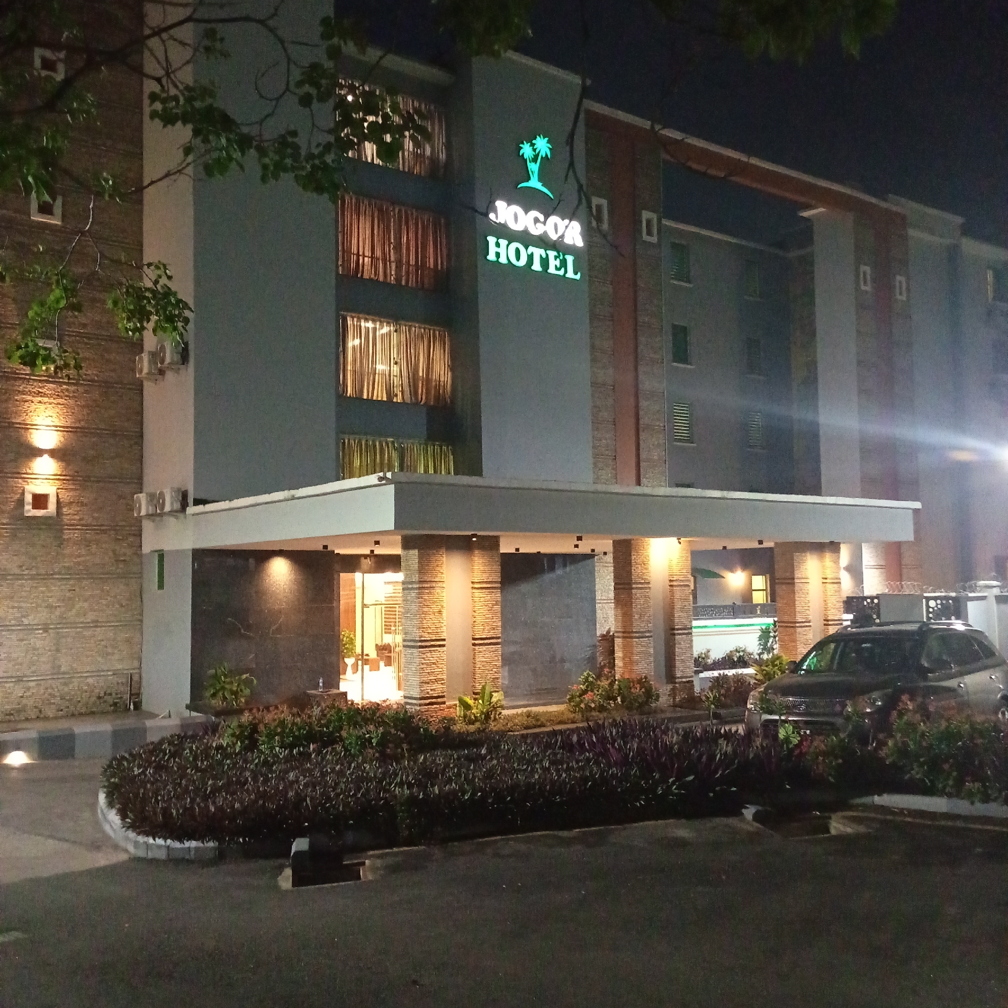An exterior night shot of the Jogor Hotel in Ibadan, Nigeria. The hotel features a modern facade with a prominent "Jogor Hotel" sign illuminated in green neon. The entrance is covered by a large canopy supported by brick pillars, and the surrounding area is landscaped with dark foliage. A dark-colored SUV is parked in front of the hotel, partially obscured by the bushes. The lighting is primarily from the hotel's internal lights, the neon sign, and some exterior lamps, creating a warm and inviting ambiance despite the darkness. The building has multiple stories with visible windows, some of which are covered by patterned curtains, suggesting the hotel is occupied. The image captures the hotel's architectural style and its presence within the urban environment of Ibadan at night.