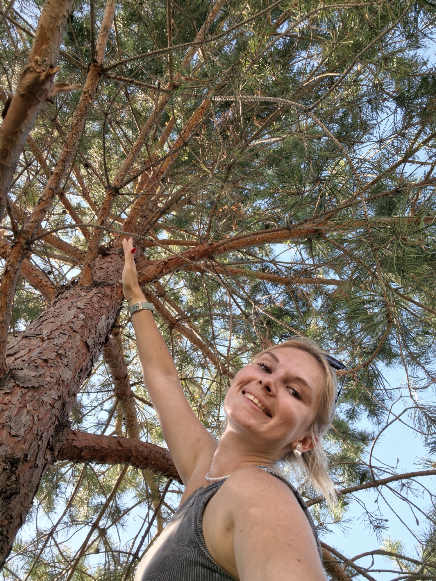 A young woman with blonde hair, wearing a grey tank top and a pearl necklace, is looking up at a pine tree. She is smiling and reaching up to touch a branch. The image is taken from a low angle, looking up through the branches of the tree towards the blue sky. There are no specific identifying features that would indicate the location is Sterlitamak, Russia.