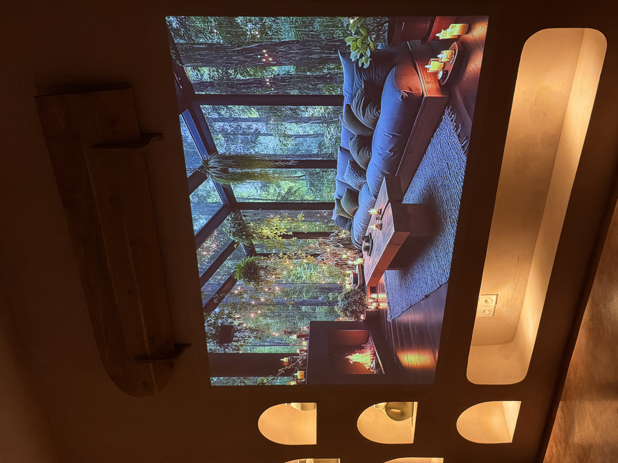A photograph depicts a wall with decorative elements. On the left, a wooden shelf is attached to the wall. To the right of the shelf, an image is projected onto the wall, portraying an interior space with a sofa, a fireplace, and lush greenery, suggesting a room with a view into a forest. The projected image shows a modern, upscale design. Further to the right on the wall is a vertical recessed design, illuminated from within, with electrical outlets visible inside the recess. Below the projected image, a series of semicircular cutouts are spaced along the bottom of the wall, also illuminated. The wall itself appears to be a neutral color. No people are visible in the image. The location, North Kuta, Indonesia, is not evident from the image.