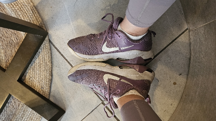 Two purple Nike sneakers with white swooshes are positioned heel-to-heel, one on top of the other, as if reflecting in a mirror. The shoes appear worn, with slightly soiled white soles and visible dirt. The person wearing the shoes is also sporting matching purple leggings and white socks. The backdrop consists of a gray stone floor with a rug and a dark brown table leg visible on the left side.
