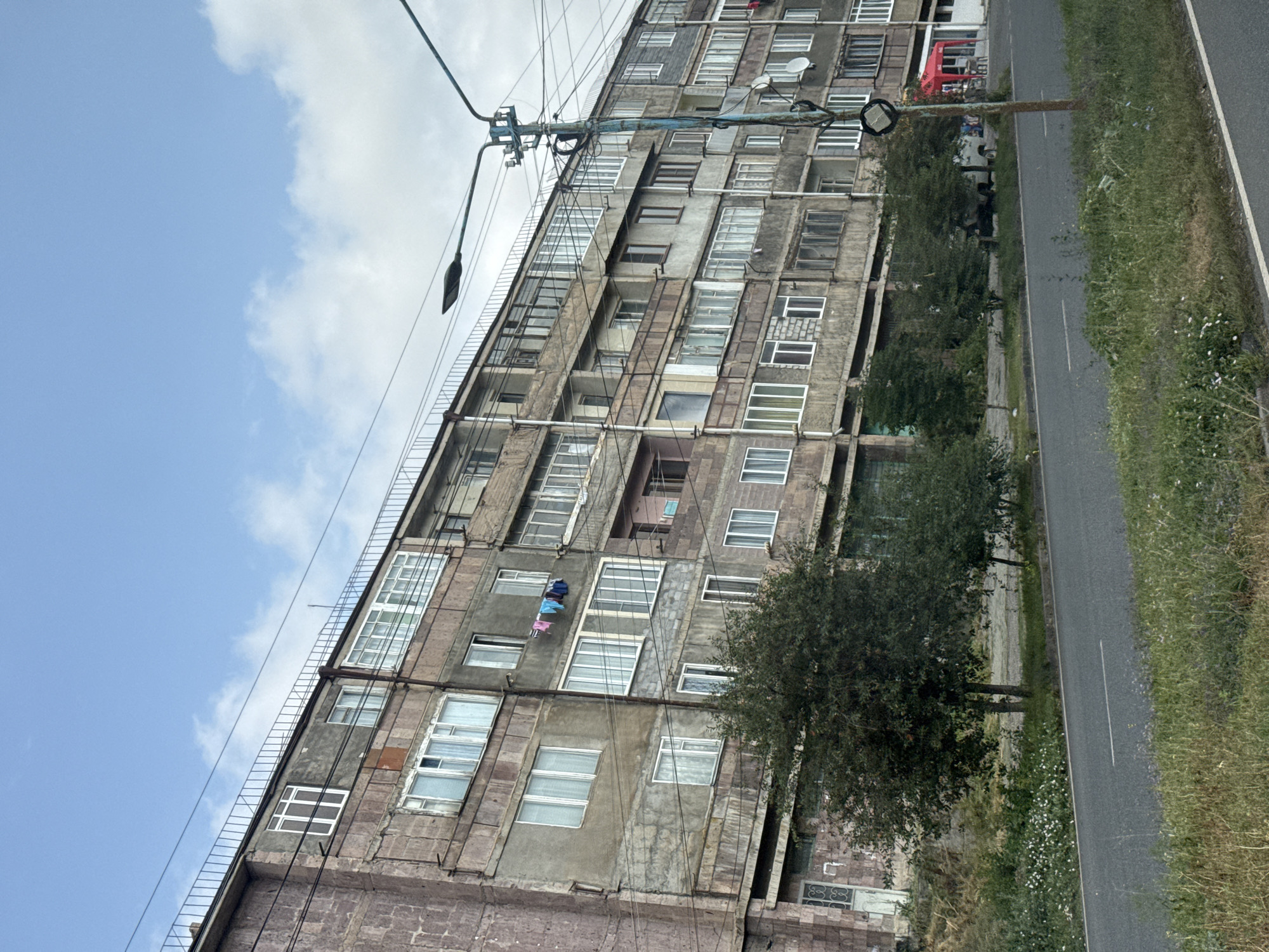 This image captures a view of an old, multi-story apartment building on a street in Sevan, Armenia. The building appears to be made of concrete and stone, with numerous windows, some of which are boarded up or appear damaged. Laundry is hanging from several balconies. There are trees and vegetation along the side of the road in front of the building. A street lamp stands on the right side of the image, and a red tent or canopy is visible in the distance. The sky is partly cloudy. The overall scene suggests an aging urban environment.