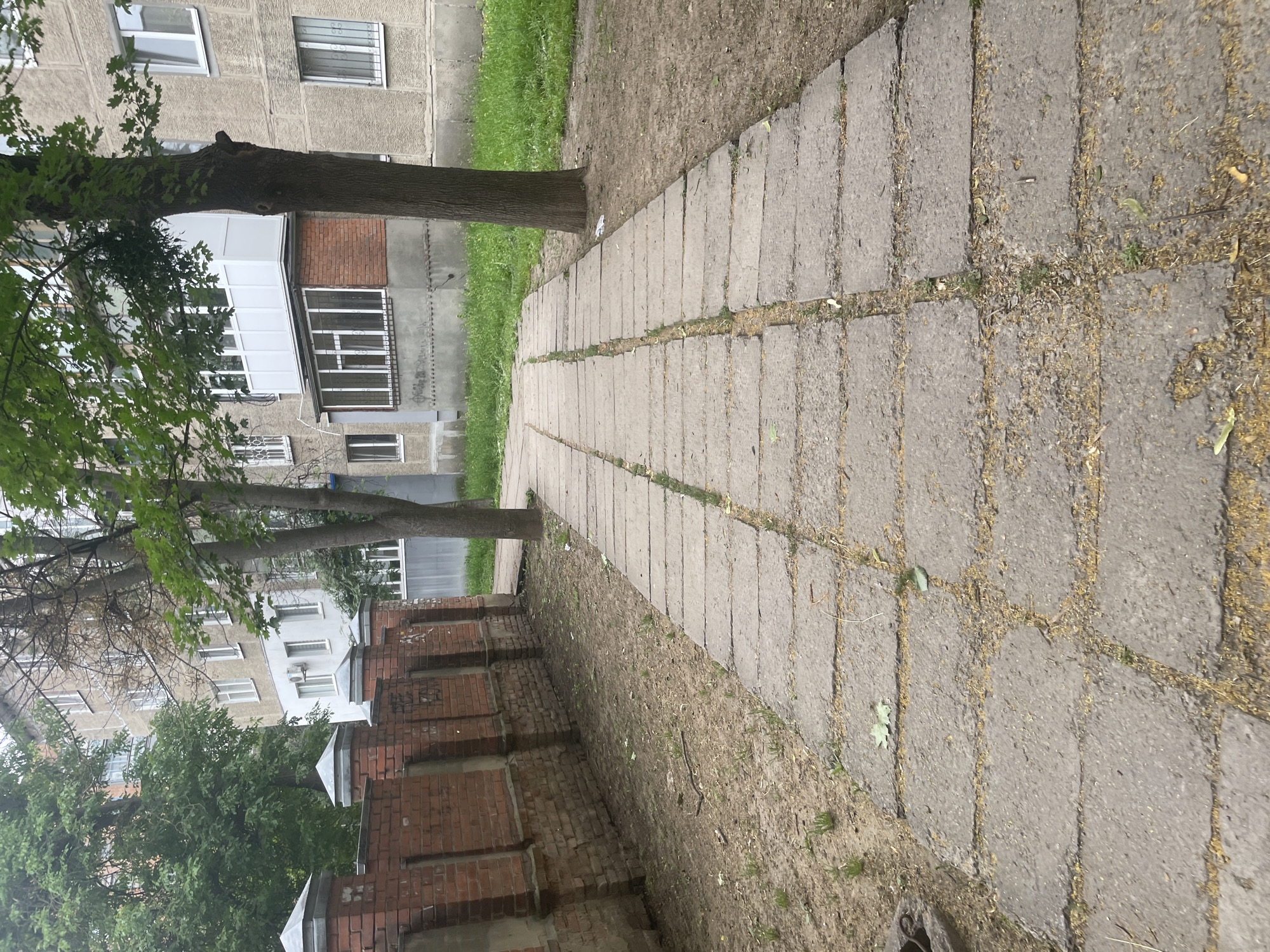 The image shows a concrete pathway made of rectangular slabs, curving towards the right. The pathway is bordered by a brick wall on the left and grass and trees on the right. A multi-story building, likely an apartment complex, is visible in the background behind the trees. The sky is overcast, suggesting a cloudy day. The overall scene depicts a residential area with a well-trodden path.