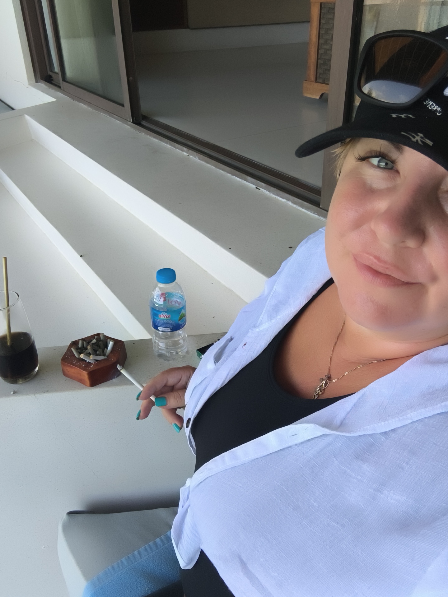 A woman with blonde hair and blue eyes is taking a selfie while on a balcony, possibly in Phu Quoc, Vietnam. She is wearing a white, unbuttoned shirt over a black top, a black baseball cap with a design, and sunglasses. Her right hand is holding a lit cigarette, and her left hand, with turquoise nail polish, is resting on her leg. In front of her, on a white ledge, is a glass with a straw, an ashtray filled with cigarette butts, and a bottle of water. The background shows a sliding glass door with a view of an indoor space with rattan furniture. The balcony has white stairs.