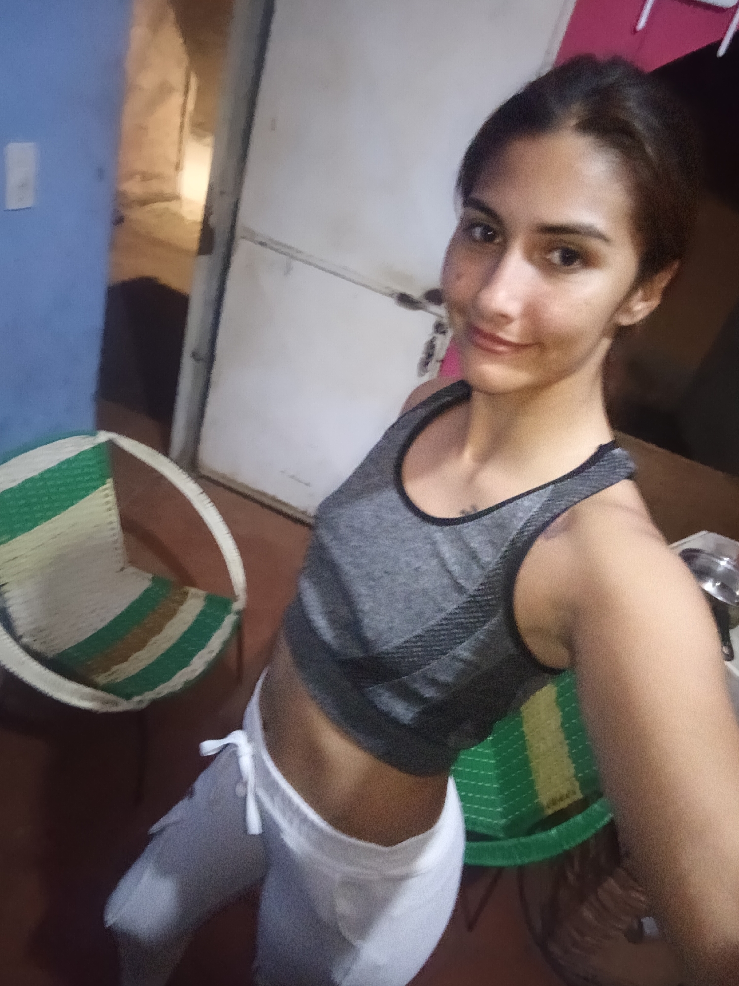 A young woman, likely in her late teens or early twenties, takes a selfie indoors in what appears to be a home in Juan Griego, Venezuela. She has dark hair pulled back, light brown skin, and is looking directly at the camera with a subtle, pleasant smile. She is wearing a grey sports bra or crop top with black trim and white drawstring sweatpants.
To her left, a round-backed chair with green and beige/white woven stripes is visible. The background features a blue wall section with a white light switch, an open doorway leading to a dimly lit interior, and a white door or wall on the right. Partially visible to her right is a green and yellow woven basket or container, and a metal pot. The lighting suggests an indoor setting, possibly under artificial light or during the late afternoon/evening. No visible text is present in the image.