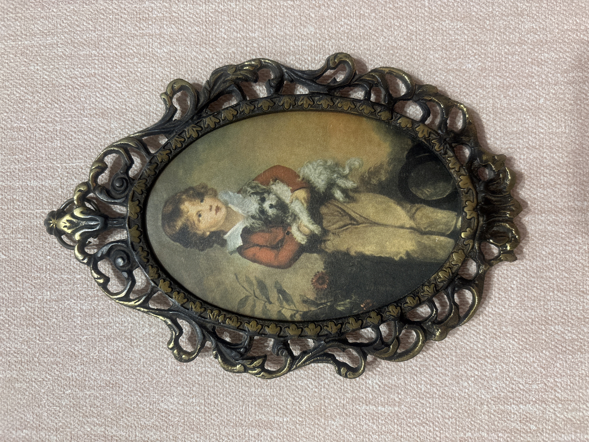 The media file shows an oval-shaped framed painting. The painting depicts a young boy dressed in historical clothing, possibly from the 18th century, wearing a reddish-brown jacket, a white collar, and breeches. He is holding a small, white, fluffy dog in his arms. The boy's expression is calm and gentle. He appears to be reclining or sitting in a naturalistic setting, with hints of foliage and a dark object, possibly a hat, in the background. The lighting suggests an indoor scene with soft, diffused light. The frame is ornate, made of metal with a dark finish and gold accents, featuring decorative scrollwork and leaf motifs. The background of the image is a light pink textured surface. There is no visible text in the image. The subject matter and style of the painting suggest a portrait from a historical period.