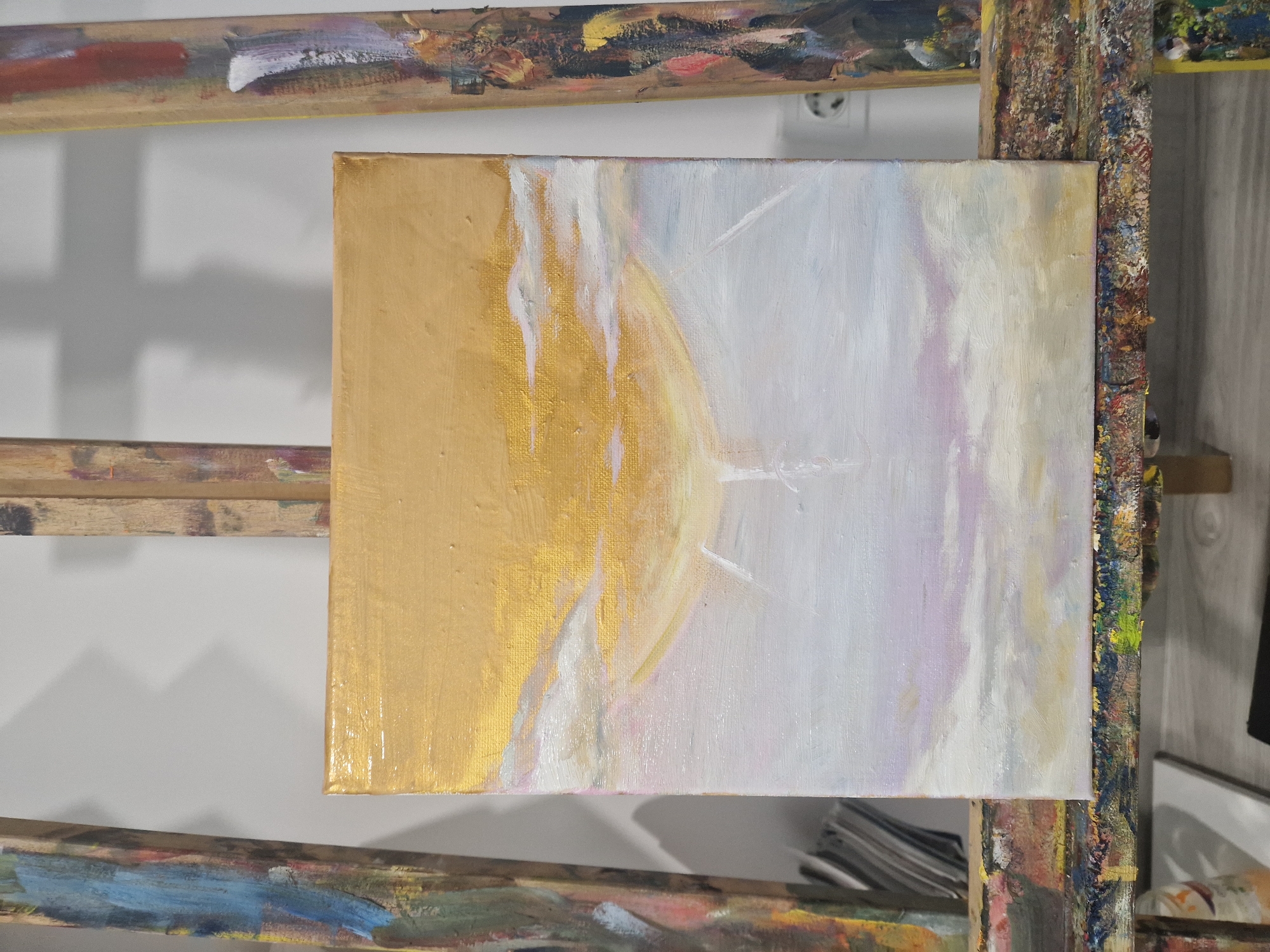 The image shows an oil painting on a square canvas, resting on a paint-splattered wooden easel. The painting is split roughly in half, with the left side featuring a golden-yellow area that may represent a sun or celestial body. The right side is a blend of soft pastel colors, mainly white, light purple, and hints of yellow, suggesting clouds or sky. Thin strokes of white paint are visible across both sections, adding texture and highlighting potential movement.
The easel itself displays a history of artistic use with layers of dried paint. The background is a plain white wall. The location is not discernible from the image. There are no people visible in the image.
