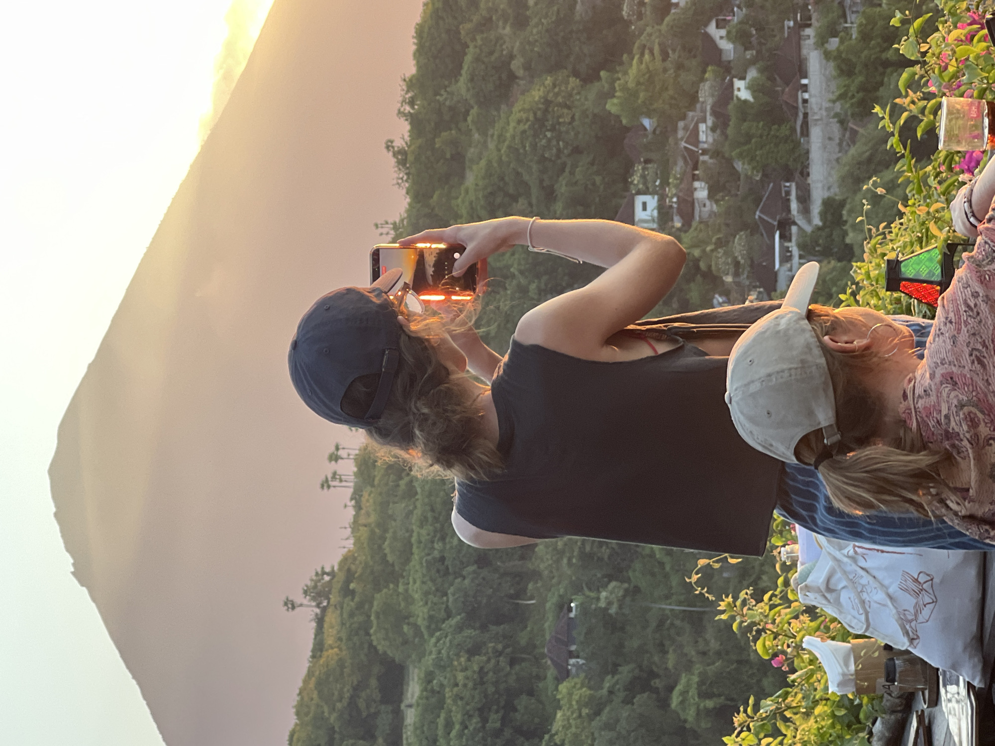 This media file captures two individuals at an overlook with a scenic view. The person in the foreground, wearing a dark t-shirt and a dark baseball cap, is holding up a smartphone to take a picture or video of the landscape. Their arm is bent, and their focus is on the device. To their right, another person is visible, wearing a light-colored baseball cap and a patterned top. They are partially obscured, with their head and shoulders visible.
The scene behind the subjects is a lush, green hillside dotted with houses, suggesting a village nestled among the trees. In the background, a large mountain is silhouetted against a hazy sky, bathed in the warm light of what appears to be sunset or sunrise. The golden hues of the sky and the soft light on the mountain indicate it is either early morning or late afternoon.
The overall mood is serene and appreciative of the natural beauty. The presence of flowering plants with pink blossoms in the foreground adds a touch of vibrancy to the scene. The location appears to be a vantage point offering a panoramic view, possibly a tourist spot or a restaurant with a view, given the presence of what looks like a table setting with a glass and some containers on the right side. There is no visible text in the image.