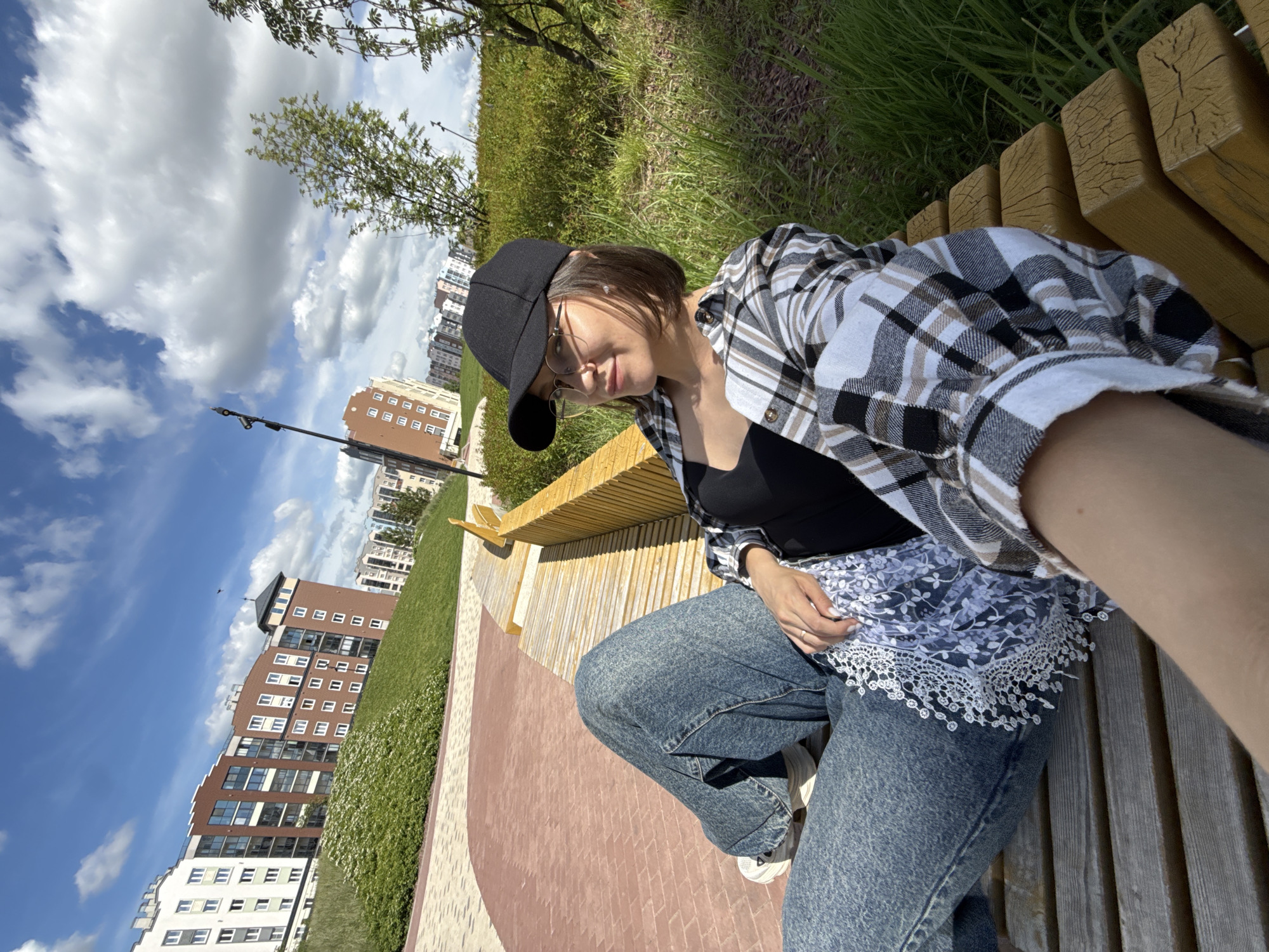 A young person is seated on a light-colored wooden park bench, captured in a selfie-style photograph. The subject has short brown hair, wears a black baseball cap, clear-rimmed glasses, a black top, a plaid flannel shirt (white, black, and beige), and blue jeans with white sneakers. A white and blue lace-patterned fabric is visible around her waist. She appears relaxed, looking towards the camera with a soft expression.
The setting is an outdoor urban park or green space. In the foreground, part of a paved area with geometric patterns in shades of brown and reddish-brown is visible. Beyond the bench and paved path, there are areas of lush green grass and bushes. In the background, several multi-story modern buildings with diverse architectural styles, including red-brick and white facades with numerous windows, rise against the skyline. Trees with green foliage are scattered throughout the scene. The sky is bright blue with scattered white, fluffy clouds. The sun is shining brightly, indicating daytime, with clear shadows visible. There is no visible text in the image.