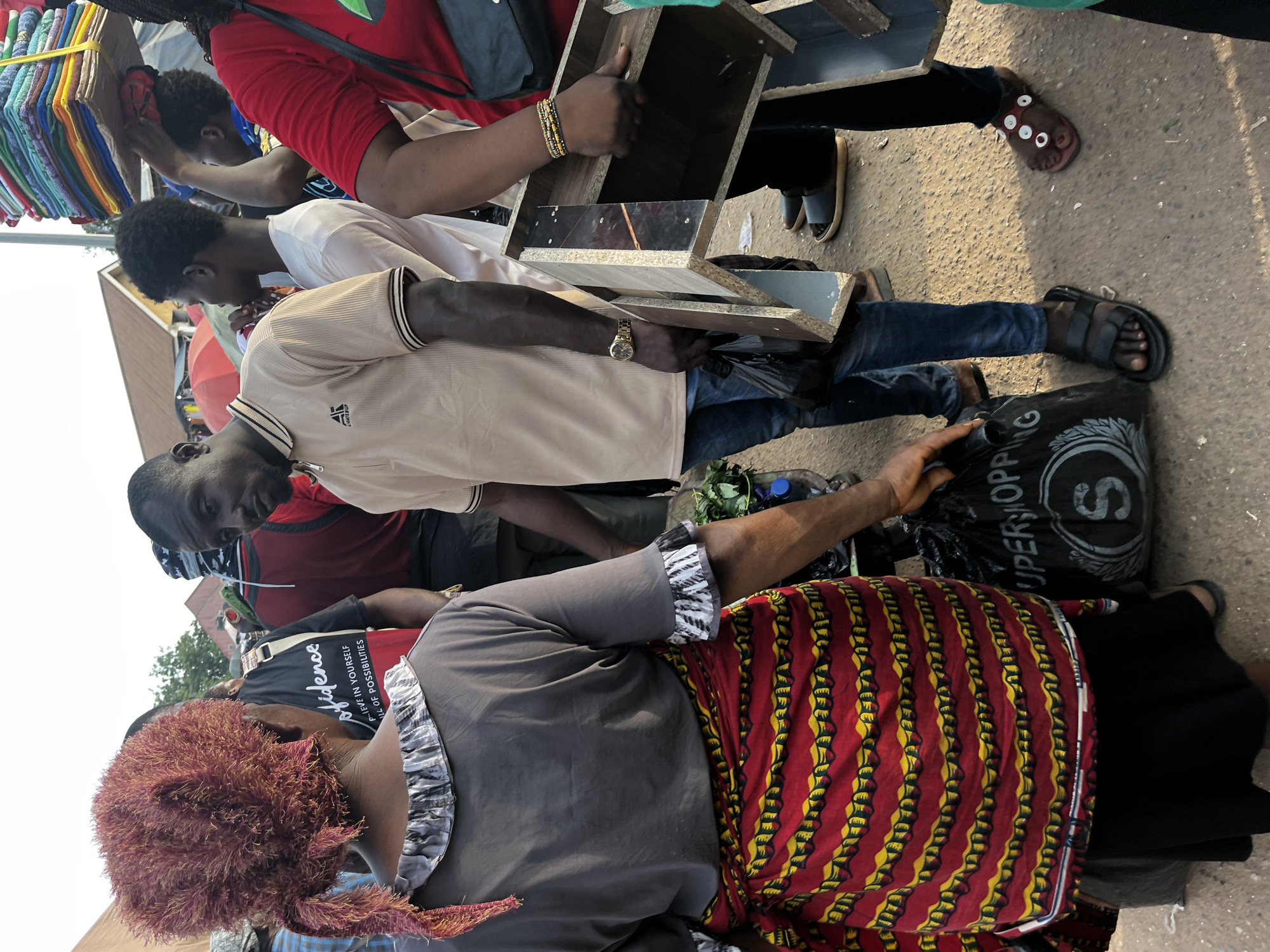 In a bustling outdoor market, likely in Nyanya, Nigeria, a group of people are engaged in commerce. The scene is vibrant and suggests a busy daytime setting, possibly a market day. The ground appears to be paved, characteristic of urban street markets.
The primary subjects are several individuals, including men and women. One person, wearing a red shirt, is partially visible on the left, with a stack of colorful textiles behind them. Another individual in a light-colored shirt is holding a wooden shelf unit with mirrored sections. A third person, wearing a black shirt with white lettering, is also present. A woman in the foreground, wearing a grey top and a vibrantly patterned red and yellow cloth, is looking down.
The activity appears to be the buying and selling of goods, with the shelf unit being a key object in focus, possibly for sale or being moved. The presence of textiles and the general market atmosphere indicate a commercial transaction.
Notable details include the gold watch worn by the person in the light-colored shirt, the beaded bracelet on the wrist of the person holding the shelf, and the "SUPERSHOP" text visible on a black bag. The weather seems to be bright, casting shadows, suggesting daylight. The overall impression is one of a lively and active marketplace.