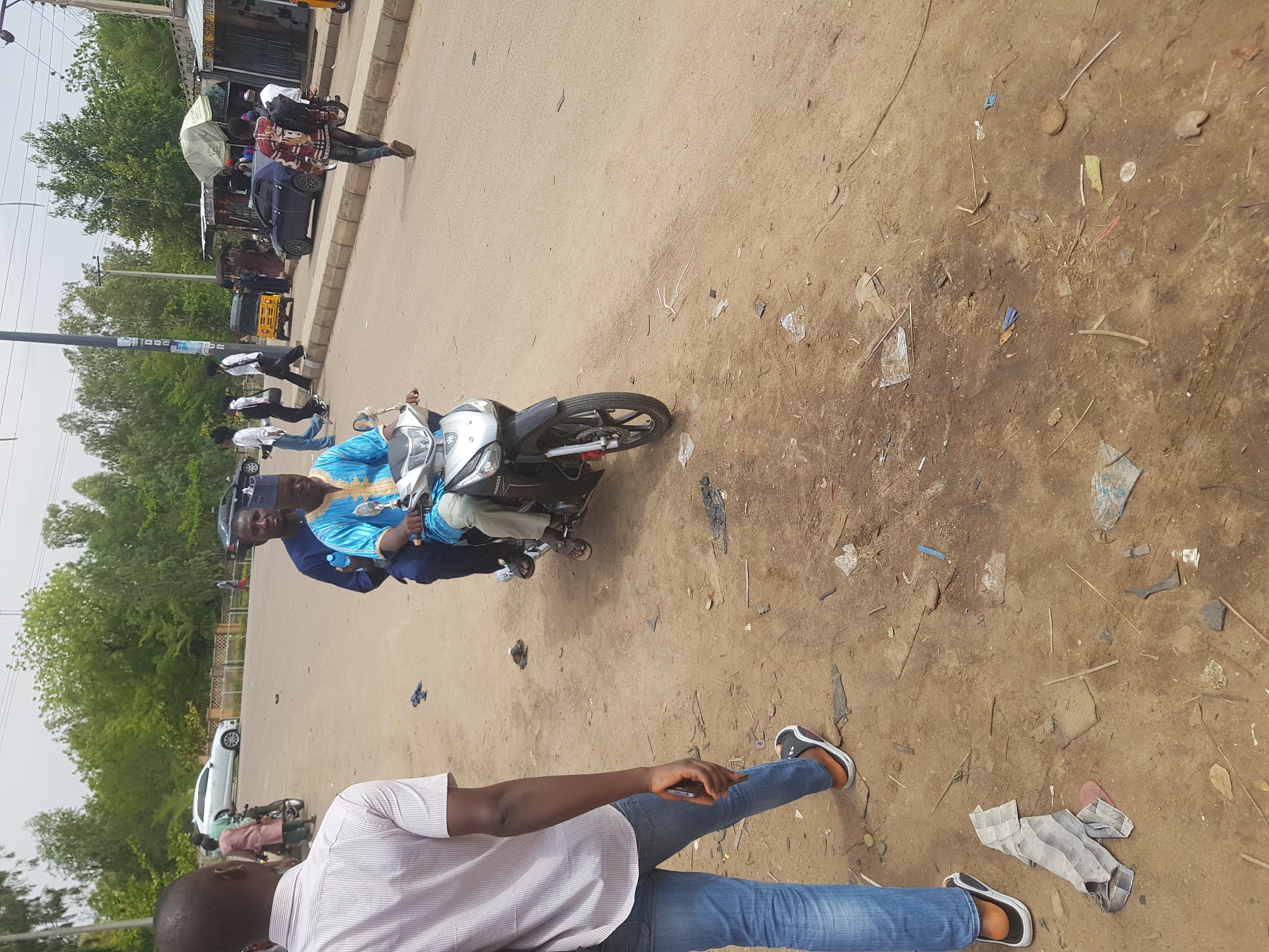The image depicts a street scene in Dabawa, Nigeria. Two men ride a black and silver motorcycle. The driver wears a blue garment, while the passenger behind him wears a dark garment. A man in blue jeans and a white striped shirt stands in the foreground, looking down at his phone. The street and surrounding dirt area are littered with debris. In the background, there are other people, vehicles, trees, and a storefront.