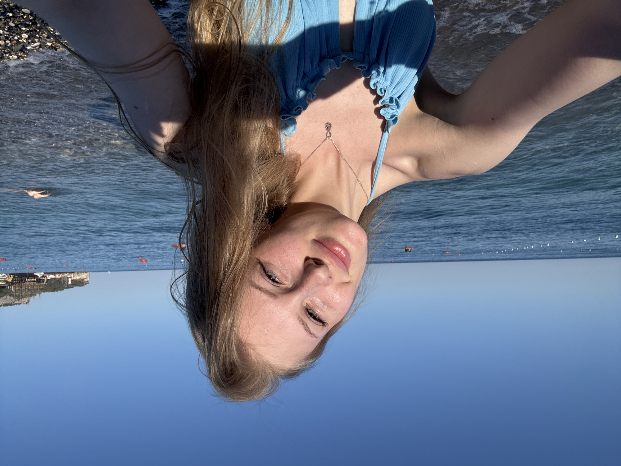 A young woman with blonde hair is taking a selfie on a pebble beach in Kemer, Turkey. She is wearing a blue bikini and a silver necklace. The sea is calm and the sky is clear blue. In the background, there are some orange buoys in the water and a hill with buildings on it. A person is swimming in the distance.