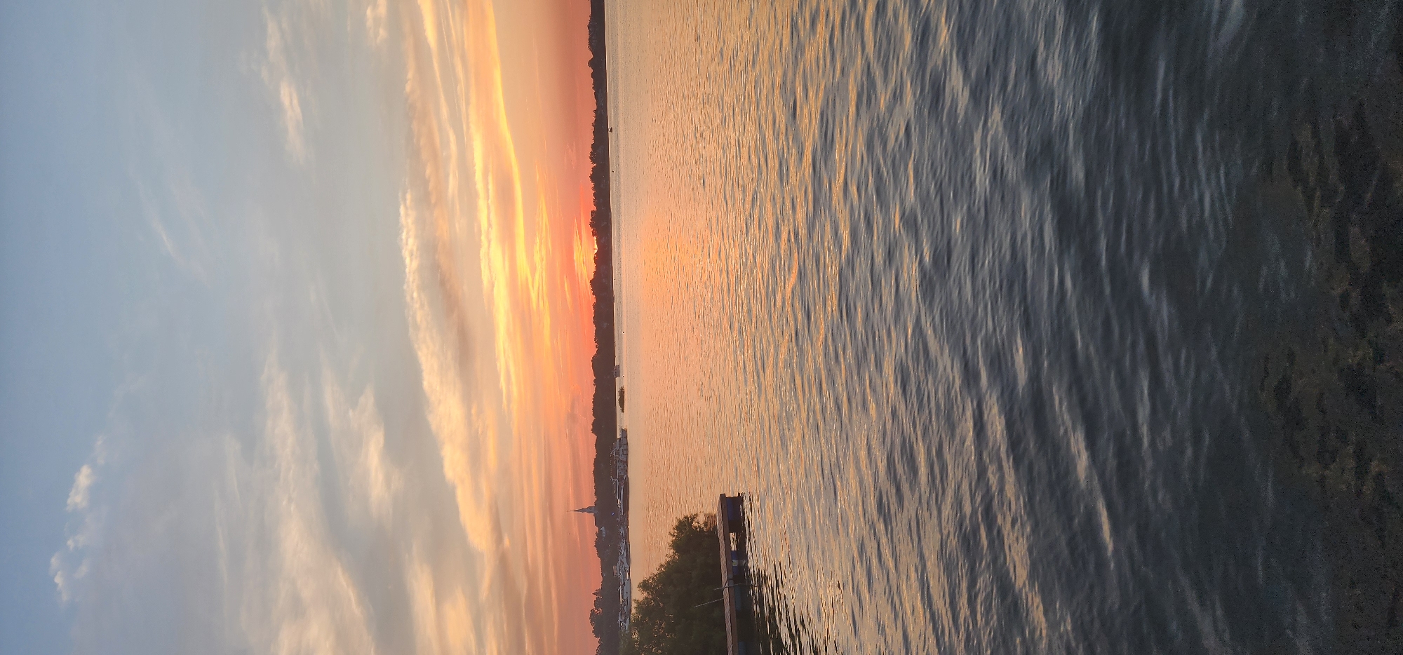 The image depicts a serene sunset over a body of water, likely a river or lake, with a distant tree line and a faint silhouette of a building with a steeple. The sky is a gradient of soft blues and oranges, with wispy clouds catching the last rays of the sun. In the foreground, the water exhibits gentle ripples, reflecting the warm hues of the sunset. To the left, a small wooden dock with dark blue floats is visible, partially obscured by lush green foliage. A few boats are scattered across the water in the mid-ground, near the shore. The overall impression is one of tranquility and natural beauty. This scene is evocative of a quiet evening on the Richelieu River in Chambly, Canada, known for its picturesque waterfront and historical landmarks like the Chambly Canal and Fort Chambly.