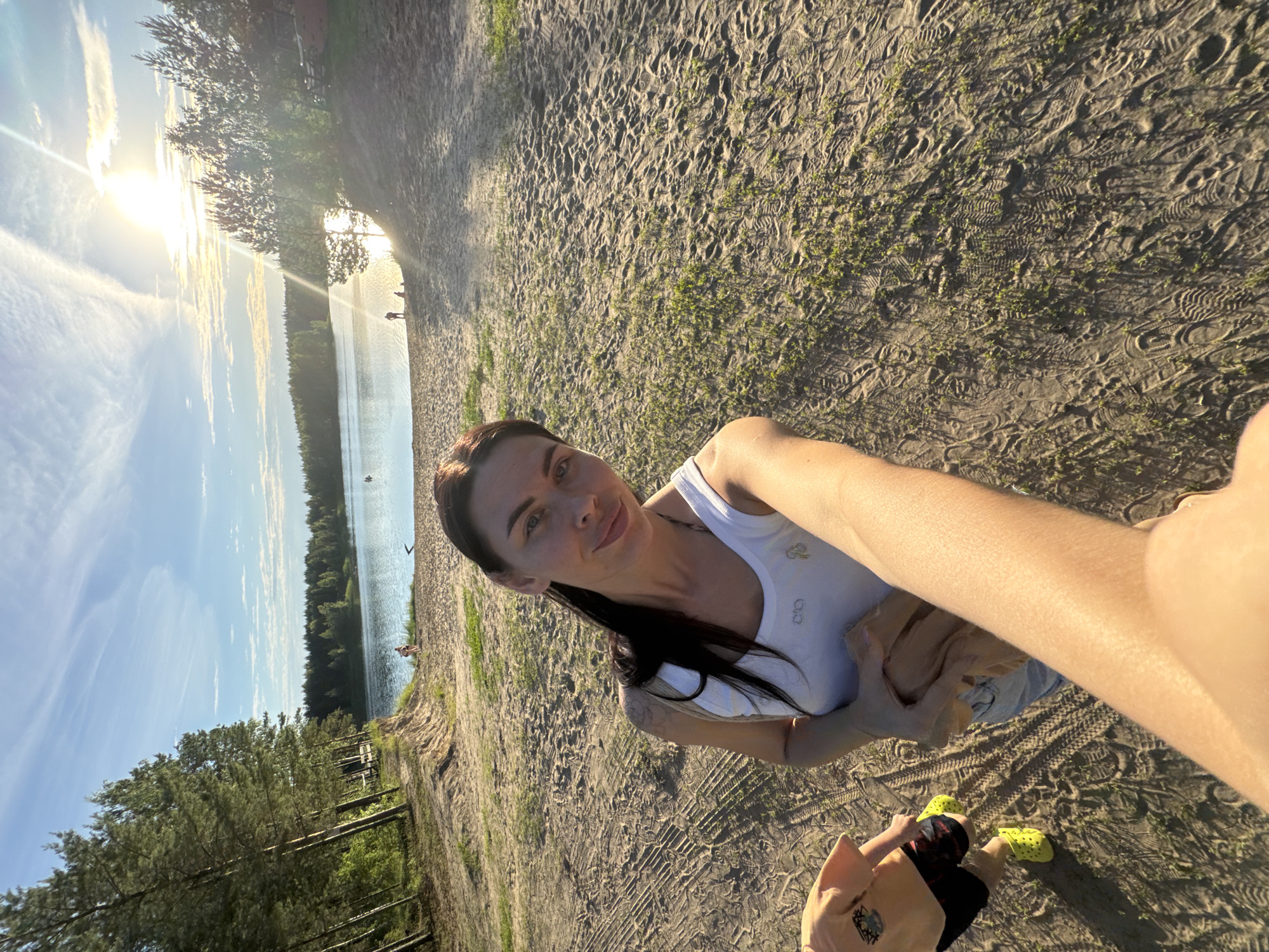 The media file shows a woman taking a selfie on a sandy beach next to a body of water. She is wearing a white tank top and has long dark hair. In the background, the sun is setting over the water, casting a warm glow. There are trees on the opposite shore and a few people are visible in the distance. The sand is marked with footprints and some small green plants are growing through it. Another person, likely a child, is visible in the lower right corner wearing a peach-colored shirt and yellow Crocs. The overall scene suggests a relaxed, outdoor setting during late afternoon or early evening. The sky is clear with some wispy clouds.