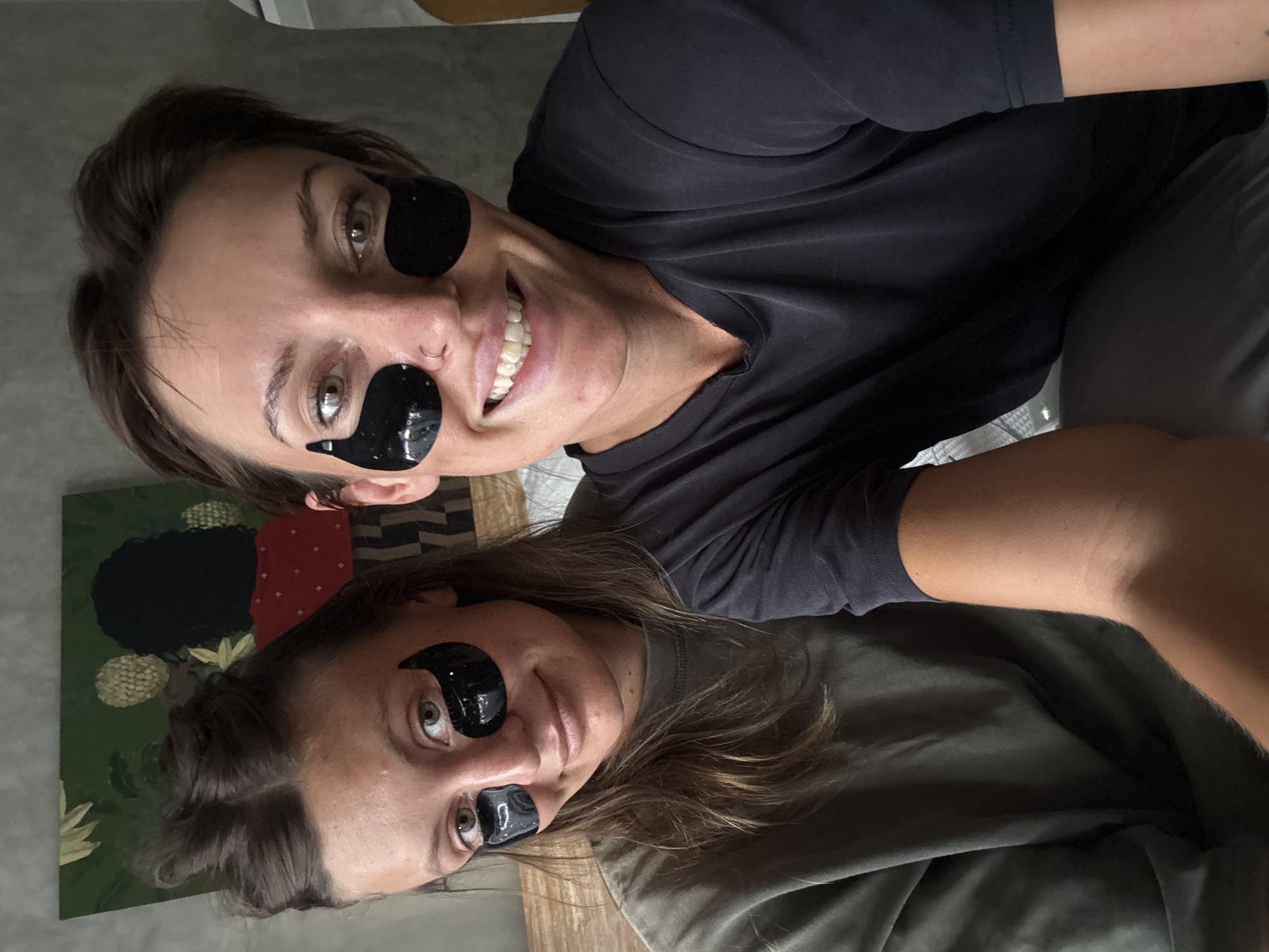 Two women are posing for a selfie while wearing black under-eye masks. The woman on top has short, dark hair and is smiling broadly, showing her teeth. She is wearing a dark t-shirt. The woman below has longer brown hair and is wearing a green t-shirt. Both women are positioned indoors. A painting with abstract floral and geometric shapes is visible in the background. The photo was taken in North Kuta, Indonesia.