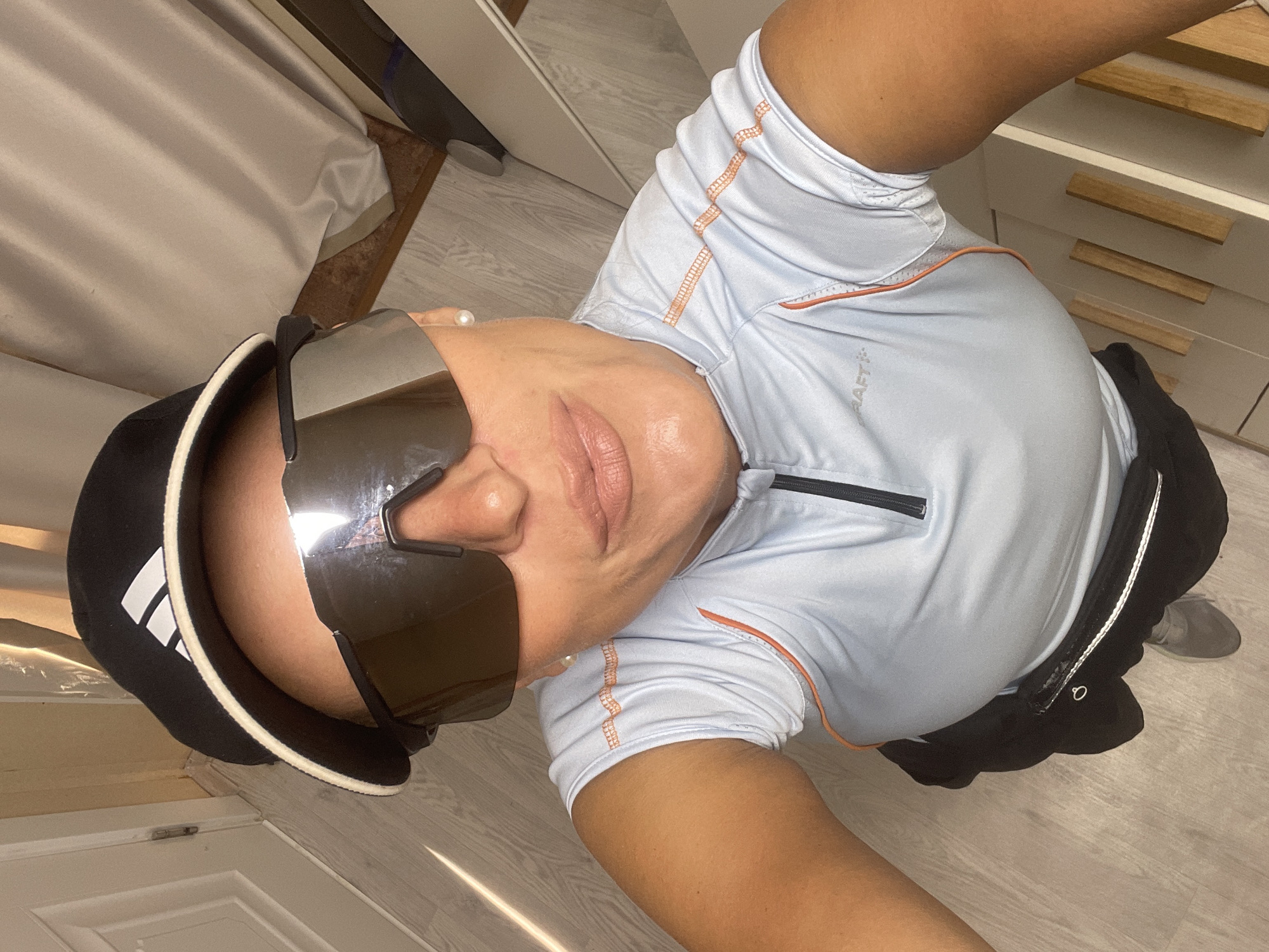 An adult is captured in an indoor, high-angle shot, possibly a selfie. The person, with tanned skin, is wearing large, mirrored, wraparound sunglasses that obscure their eyes, and a black cap with a white brim and a visible white Adidas logo on the side. Pearl-like earrings are visible on both ears. The person is dressed in light blue athletic wear with contrasting orange stitching and a black zipper on the chest, bearing the visible text "CRAFT:". They are also wearing black athletic shorts or leggings with a white stripe down the side. The expression is unreadable due to the sunglasses, but the pose appears confident.
The background features a light-colored wooden floor, a gathered light-colored curtain or fabric on the left, and white furniture with wooden handles on the right. A grey shoe is partially visible on the floor to the right. The lighting suggests an indoor setting with ambient light. The scene is located in Baranovka, Russia.