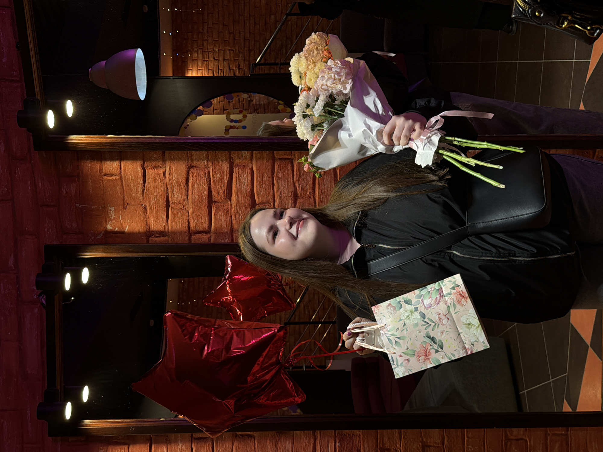 A young woman with long, straight brown hair smiles at the camera, holding a large bouquet of flowers and a gift bag. She is wearing a black jacket and a dark shoulder bag across her body. The bouquet, wrapped in white paper and tied with a pink ribbon, features various light-colored blooms, including white, cream, pink, and orange flowers like chrysanthemums and roses. The gift bag has a floral pattern with pink, green, and white flowers on a light background.
The setting is an indoor space, likely a restaurant or event venue, characterized by reddish-brown brick walls and dim, atmospheric lighting. A large mirror or reflective surface behind the woman shows reflections, including her own face and two shiny red star-shaped balloons. Several small spotlights illuminate the scene, mounted on the mirror's frame, and a larger, dark pendant light is visible in the upper background. The dim lighting suggests it is evening or night. The woman's joyful expression indicates a celebratory mood. A partial reflection in the mirror reveals what looks like a decorative sign with colorful circles and possibly the number "10" or the Cyrillic letter "Ю". The overall scene captures a moment of receiving gifts and celebrating.