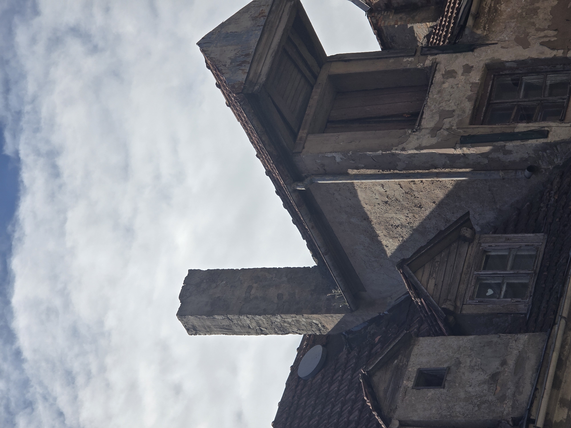 A low-angle, close-up, eye-level shot captures the weathered facades of old buildings against a partly cloudy sky in Riga, Latvia. In the foreground, the tiled roof of a building features a square, dark satellite dish. To the right of the dish is a tall, rectangular chimney made of rough, grey stone blocks. Beyond the chimney, another building's roof is visible, with a prominent, angular extension jutting out. The plasterwork on these structures is heavily degraded, with peeling paint and visible brickwork. Two dormer windows are present on the left side of the image. The left dormer has a small, dark, square window with a visible pane. The right dormer, constructed with wooden planks, features a larger window divided into two panes, with a small white heart shape visible on the interior of the glass. A drainpipe runs vertically along the side of one of the buildings. The overall scene depicts urban decay and neglect.