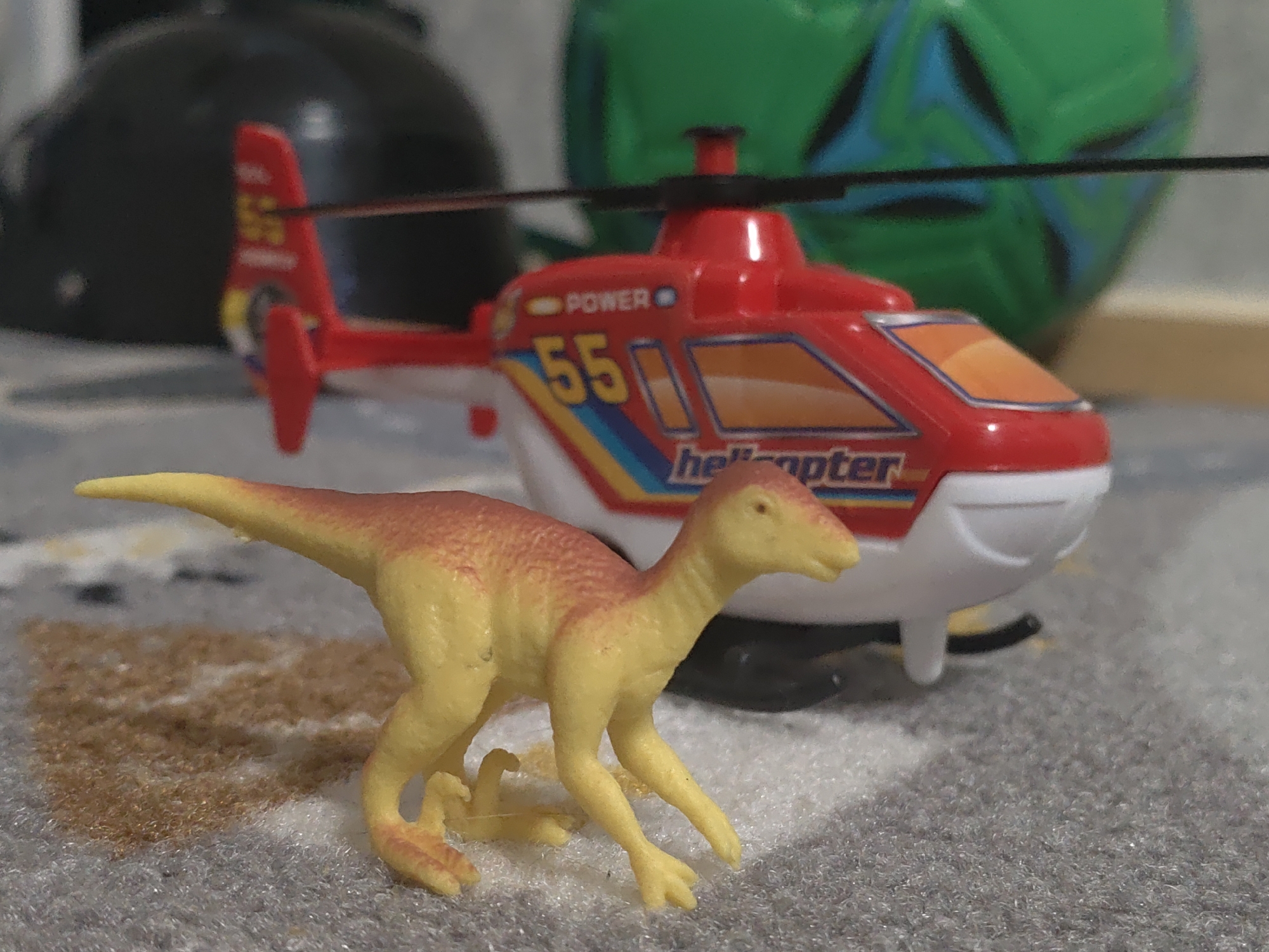 A close-up, low-angle, eye-level outdoor shot depicts two toys, a yellow and orange dinosaur figurine in the foreground and a red toy helicopter in the background, positioned on a grey, textured surface that resembles carpet. The dinosaur is facing right, with its body slightly angled, and its right leg is bent, suggesting movement. The helicopter, prominently branded with "POWER" and the number "55," has a white fuselage and red accents. The environment is indistinct, with a blurry black object on the left and a green, circular object with blue markings on the right, also out of focus. The image does not contain any identifiable landmarks, people, or elements that would allow for localization to Kurganinsk, Russia.