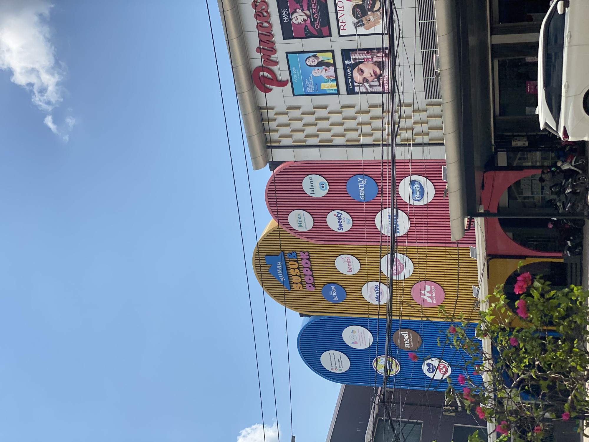 The image shows a bright blue sky with scattered white clouds above a building exterior in Banjarmasin, Indonesia. The building features a colorful facade with arched sections in blue, yellow, and pink, adorned with various circular logos for brands such as Lactogen, Bambi, Merries, Moell, Morinaga, Milna, Loluna, Sweety, and Gently. Above these, the text "RUMAH SUSU POPOK" is visible in a stylized font. To the right, a white building with gold decorative elements displays advertisements for "Princess," "Wardah," "Maybelline," and "Revlon." In the foreground, a white car is parked, and to the left, pink bougainvillea flowers bloom on a tree. The street in front of the building has several parked motorcycles, and a few individuals are visible near the entrance of the building. Power lines are visible crisscrossing the upper portion of the frame.