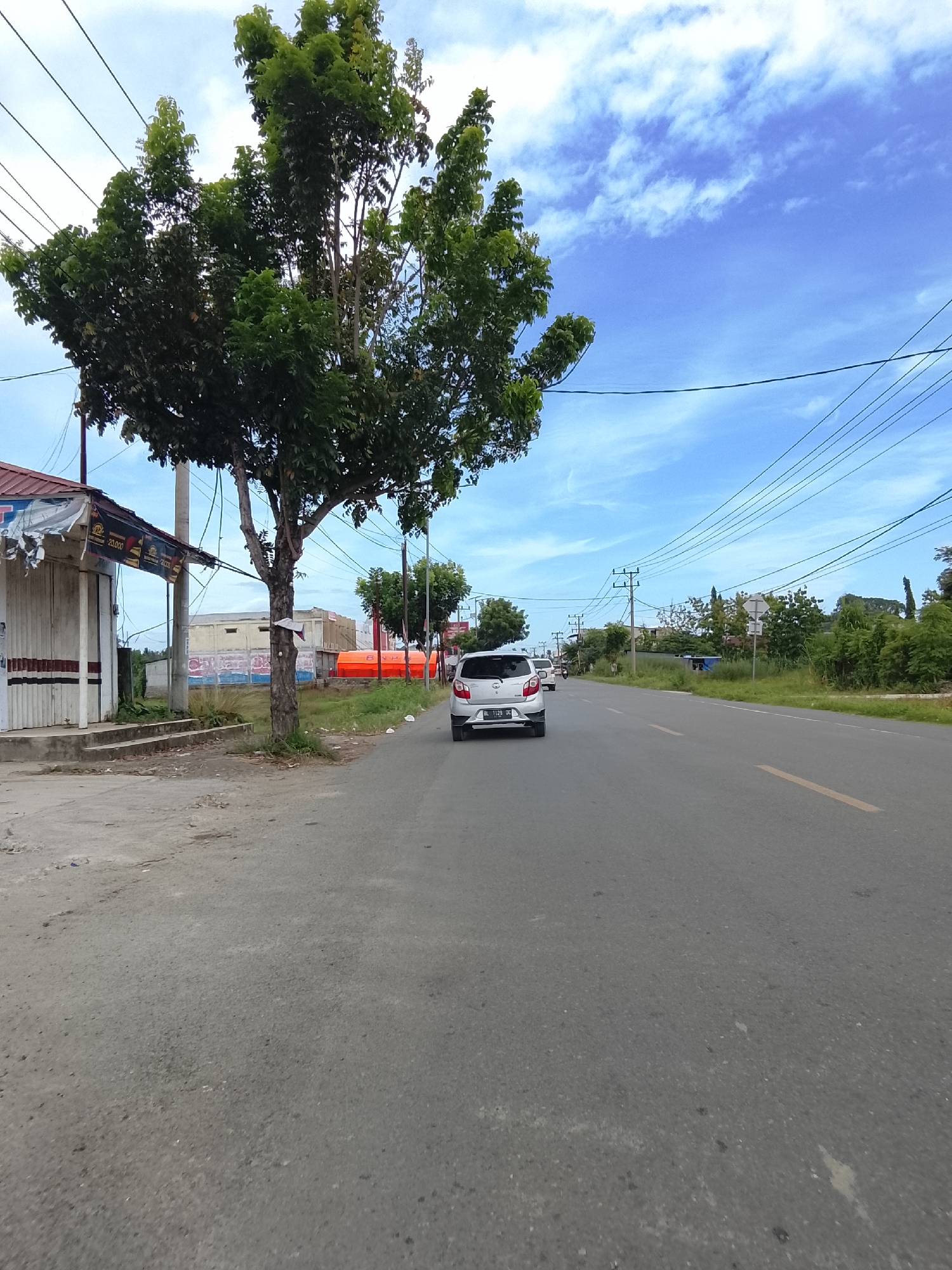 The image depicts a street scene in Bunot, Indonesia. In the foreground, a paved road with a yellow dividing line runs towards the horizon. On the left side of the road, there is a partially obscured building with a corrugated metal roof and white roll-up doors. A tree with dense green foliage stands next to the building. Beyond the building, several orange cylindrical tanks are visible. Further down the road, a silver car with a license plate reading "BL 1128 QC" is driving away from the camera. The road is lined with vegetation and utility poles with numerous overhead wires. The sky is blue with scattered white clouds. The scene appears to be a typical roadside view in a semi-urban or rural area.