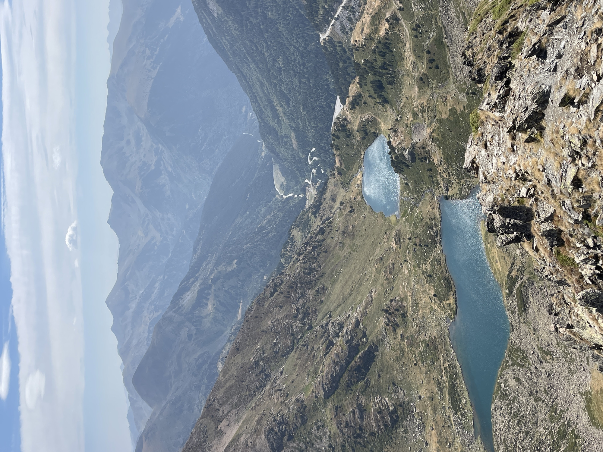 An elevated, wide-angle, sunlit view depicts a mountainous landscape with two shimmering blue lakes in the mid-ground. In the foreground, a rocky, dry, and sparsely vegetated slope descends into the valley. The mid-ground is characterized by rolling hills covered in dry grass and scattered rocks, leading to the two lakes. The first lake, positioned in the lower left, is larger and more elongated, while the second, slightly higher and to the right, is smaller and more circular. Beyond the lakes, a valley unfolds, densely forested with dark green trees on its slopes. A winding road with multiple switchbacks is visible on the right side of the valley, ascending the mountainside. The background features a series of layered mountain ranges that recede into a hazy blue distance, disappearing into a light blue sky with scattered white clouds. The overall scene is a natural, undeveloped environment.