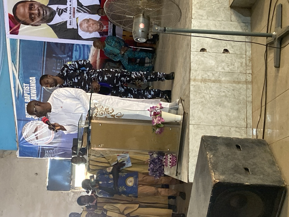 This image captures an indoor event taking place on an elevated stage within a structure in Ube, Nigeria, identified by a banner as 'Bethel Baptist Church Akwanga.' A dark-skinned male speaker, wearing glasses and a white long-sleeved traditional garment, stands centrally behind a clear acrylic and golden-based podium, holding a red-tipped microphone in his right hand. An open book is visible on the podium. To his right, a dark-skinned male in a blue/grey camouflage uniform stands upright, observing. On the stage's left, multiple individuals in light brown robes are present, one of whom, wearing a blue patterned shirt, holds a mobile phone, pointed towards the speaker. Behind the stage are large banners, one blue with a church emblem, and another displaying portraits, including 'Rev. Dr. Bulus A. Akwanga.' Two floral arrangements are positioned on the stage floor in front of the podium. Off-stage, a large black audio speaker box is in the lower left, and a grey standing fan with visible electrical cords is in the lower right. The stage features a tiled facade, and the background walls are light-colored and textured, with a bright light source visible on the far left.