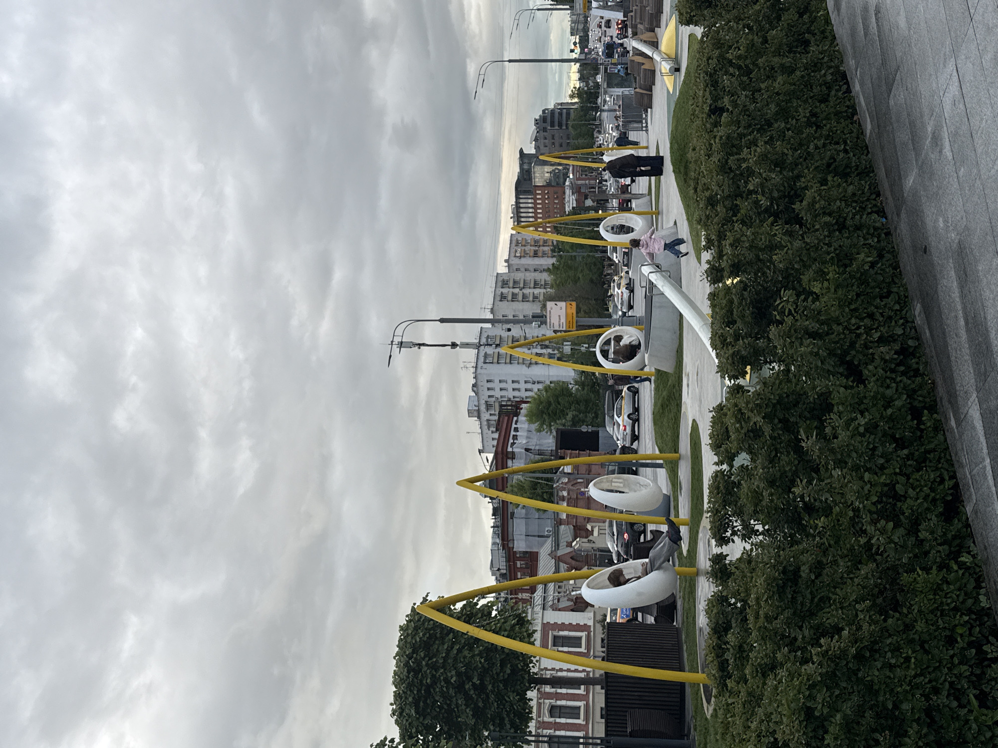 The image shows a public space in Moscow, Russia, featuring modern seating structures resembling swings with white, circular enclosures suspended by yellow frames. These structures are set along a walkway lined with greenery, including shrubs and trees. People are using the seating, with some individuals seated inside the enclosures and others walking nearby. In the background, buildings of varying heights and architectural styles are visible, characteristic of an urban environment. The sky is overcast with a uniform layer of clouds, suggesting dull or indirect lighting conditions. The scene conveys a sense of contemporary urban design intended for public relaxation and interaction. The road on the left is full of cars.