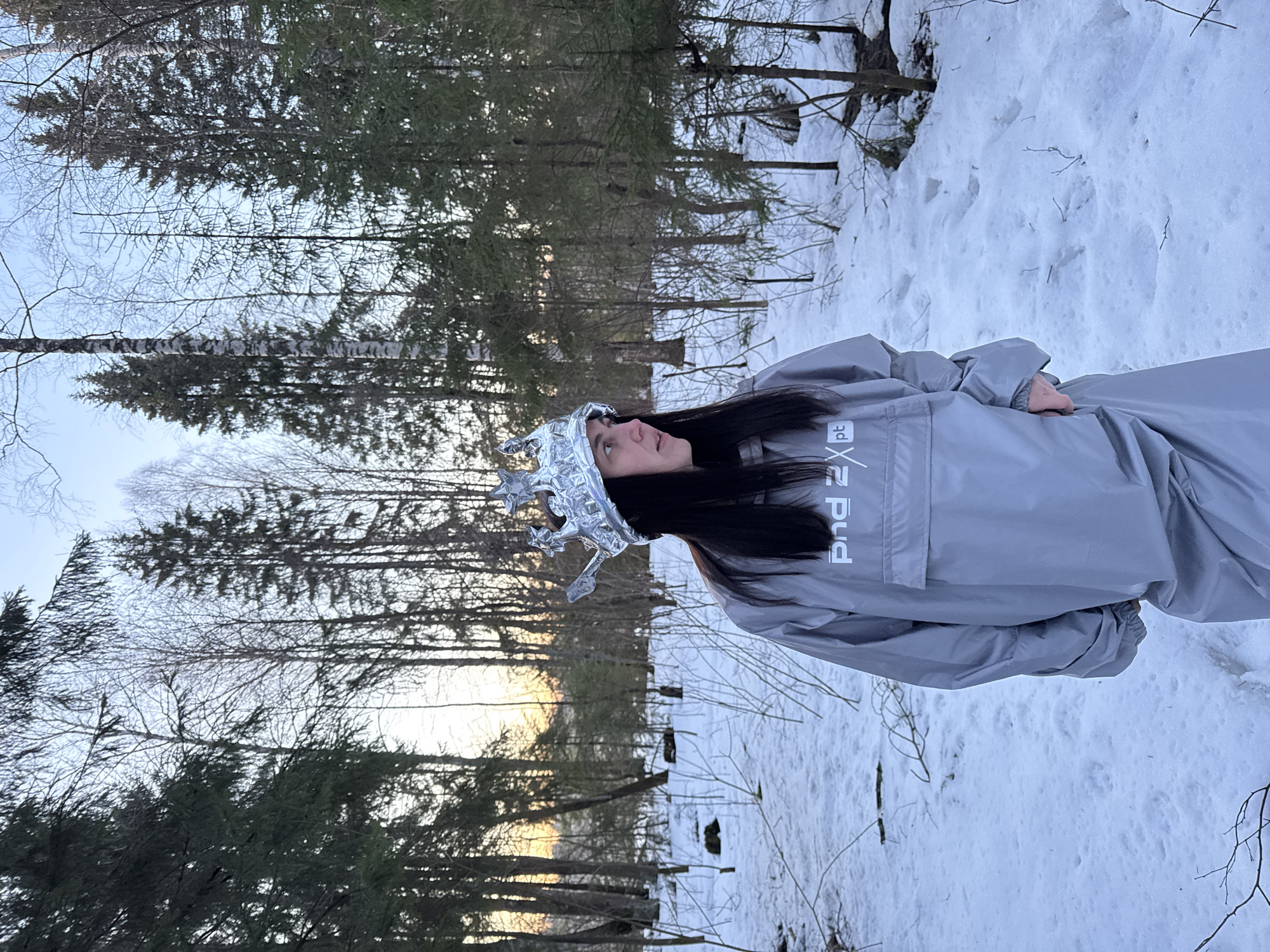 A woman stands in a snowy forest in Вассята, Russia. She wears a grey jacket with "Pnd 2 pt" printed on it, dark hair, and a foil crown. The background features tall trees, snow-covered ground, and a glimpse of the sky near the horizon.