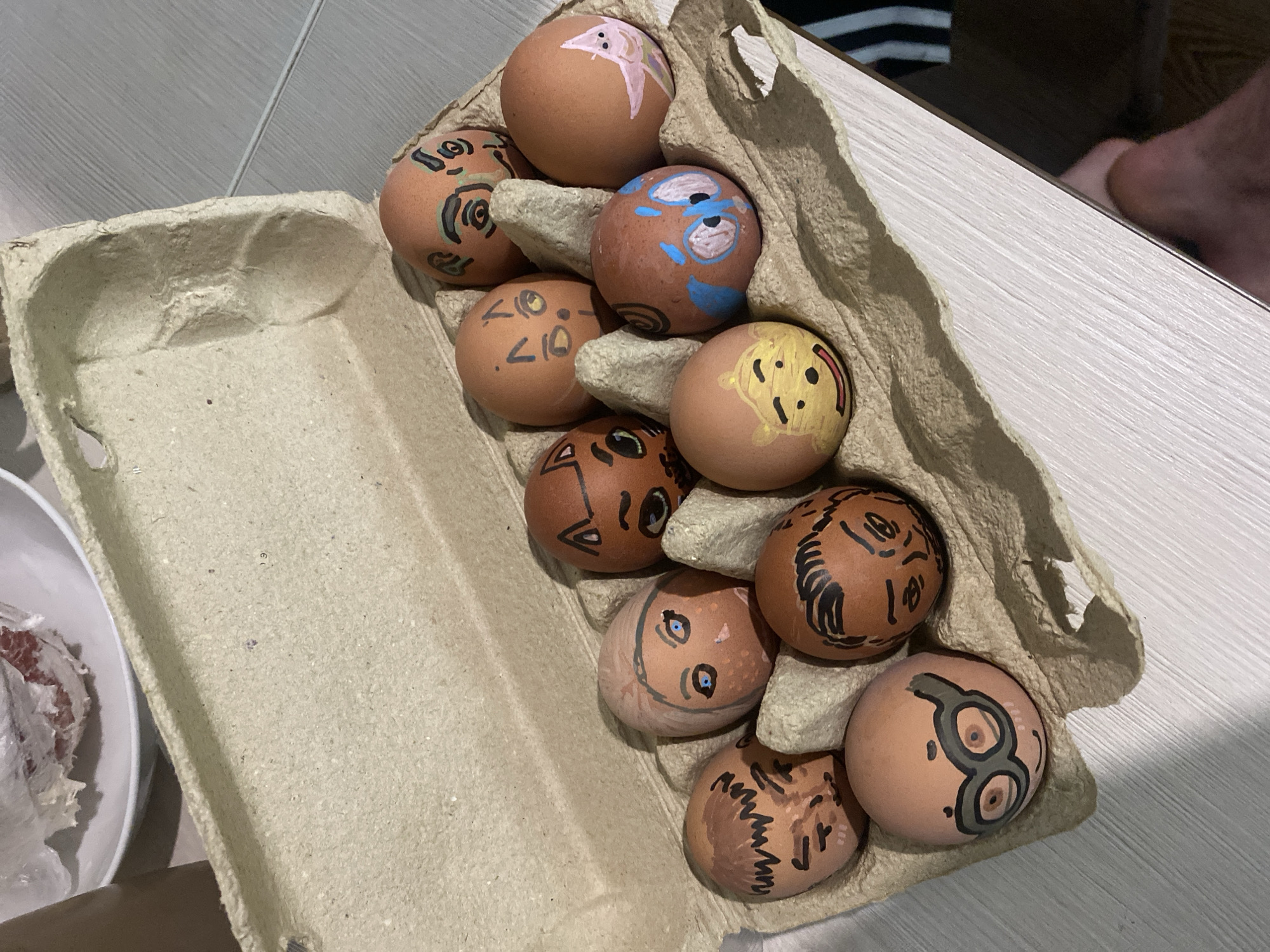 A top-down shot shows eleven brown eggs with hand-drawn faces in a tan cardboard egg carton. The eggs are arranged in the egg carton, partially filling the available slots. The egg carton sits on a light-colored, possibly laminate, tabletop. A white plate containing a white, possibly food-related, item is partially visible in the lower left corner. In the upper right, a glimpse of a person's bare foot and striped clothing is visible. There is no indication of a location in Izhevsk, Russia.