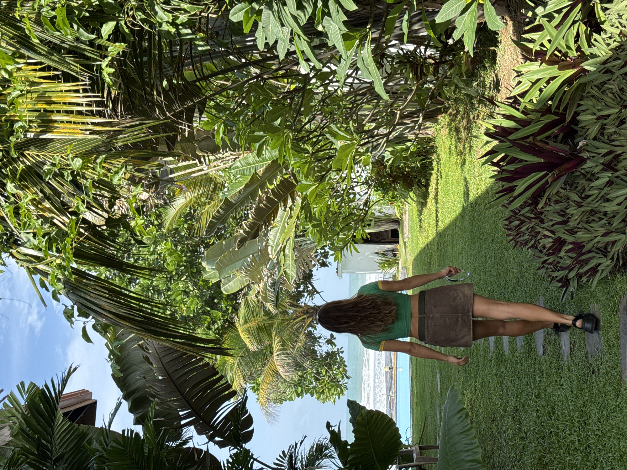In Goviyapana, Sri Lanka, a woman with long brown hair, viewed from the back, walks on a stone path through a lush garden towards the ocean. She is wearing a green crop top with yellow trim and a brown A-line skirt. She carries a pair of glasses in her right hand. The garden features manicured grass, palm trees, and various tropical plants. In the background, a bright blue swimming pool is visible, and beyond that, the ocean meets the horizon under a clear sky with scattered clouds. The scene suggests a serene tropical resort setting.