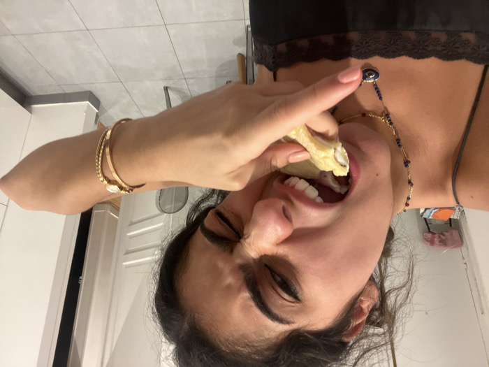 A woman with dark hair eats a filled pastry. She wears gold bracelets, a blue and gold necklace, and a black top with lace trim. Her eyes are closed, and she is holding the pastry with her right hand. The background includes a white cabinet and light-colored tiled floor. A fan is partially visible behind her arm. The woman appears to be indoors, possibly in a kitchen.