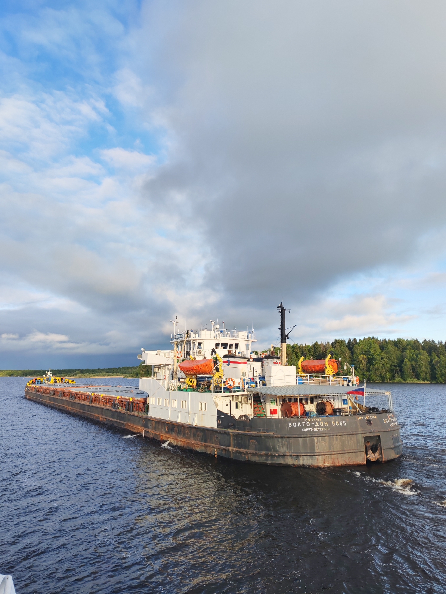 A long, wide river barge, named "ВОЛГО-ДОН 5065 САНКТ-ПЕТЕРБУРГ" and flying a Russian flag, cuts through dark water under a sky with blue patches and heavy clouds. It is likely moving from left to right in the frame. The forest lines one bank of the river.