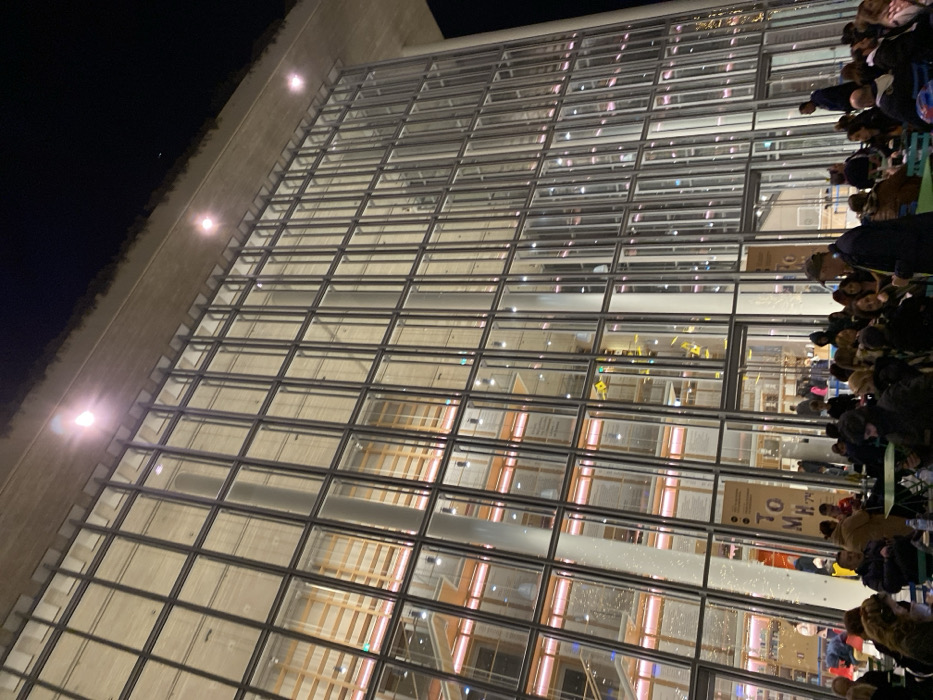 The image, captured at night and presented at a diagonal orientation, features a modern building with a prominent glass and metal grid facade, alongside a gathering of people outdoors. The building's upper structure consists of a light-toned material, from which several bright lights illuminate the facade against the dark sky. Through the expansive glass panels, internal lighting and architectural details of the building's interior are discernible.
On the right side of the frame, a crowd of people is visible, many appearing to be seated at tables on an outdoor terrace or plaza adjacent to the building. They are dressed in casual attire, suggesting a social gathering, an outdoor dining experience, or an event.
The time of day is clearly night, indicated by the dark background and the reliance on artificial lighting. A sign or poster visible inside the building through the glass facade contains partially obscured text, which includes elements such as 'TO M H'. The scene depicts a vibrant evening atmosphere at what appears to be a public or cultural venue, specifically located in Kallithea, Greece.