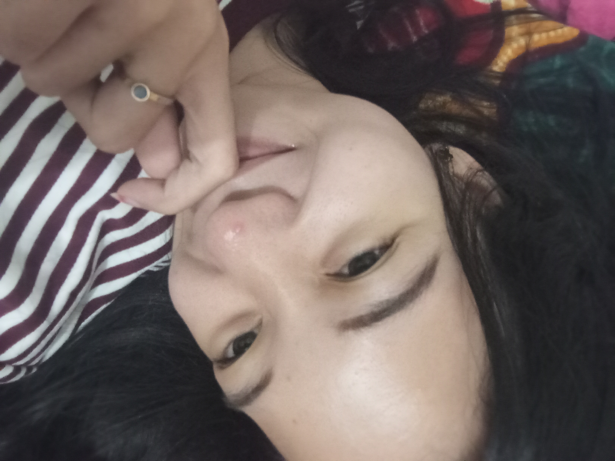 The image is a close-up selfie of a young woman lying down. She has long, dark hair and is wearing a striped shirt. Her right hand is resting on her chin, and she is wearing a ring with a dark stone on her index finger. The background appears to be a bed or couch with patterned fabric. The lighting is soft, suggesting an indoor setting. There is no discernible information about the location in Rejosari, Indonesia, within the image itself.