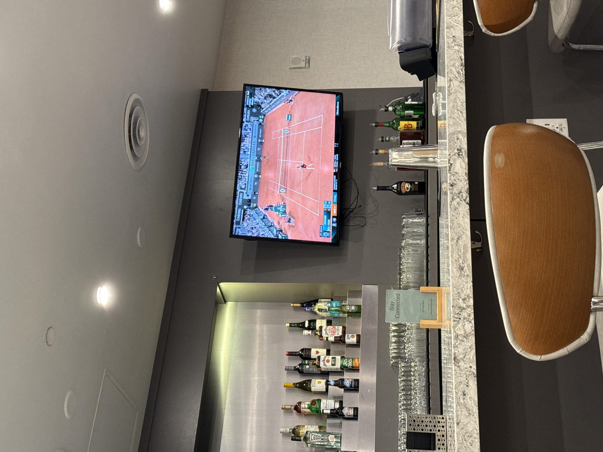 The image depicts a modern bar area, likely within an airport lounge. A large flat-screen television is mounted on a dark grey wall, displaying a tennis match on a clay court. The screen shows "BNP PARIBAS" sponsorship, "FINAL" status, player names including "SABALENKA," and game scores. "Emirates" branding is also visible on the TV screen.
Below the television, various liquor bottles are arranged on illuminated shelves, including "JIM BEAM," "BAILEY'S," and a partially visible "COORS LIGHT." A light-colored granite bar counter runs along the right side of the frame, laden with rows of clean glassware. Two modern bar stools with light wood backs and white upholstered seats are visible, pulled up to the counter.
A small green sign on the bar countertop reads "Stay Connected" and includes "The Club MCO" and "Emirates," indicating the location is likely an airport lounge at Orlando International Airport (MCO). An electrical outlet on the wall behind the stools is labeled "LMC-21." Overhead lighting fixtures and a circular air vent are visible on the light grey ceiling, contributing to the indoor, contemporary setting. The scene suggests a leisure area for travelers to relax and watch sports.