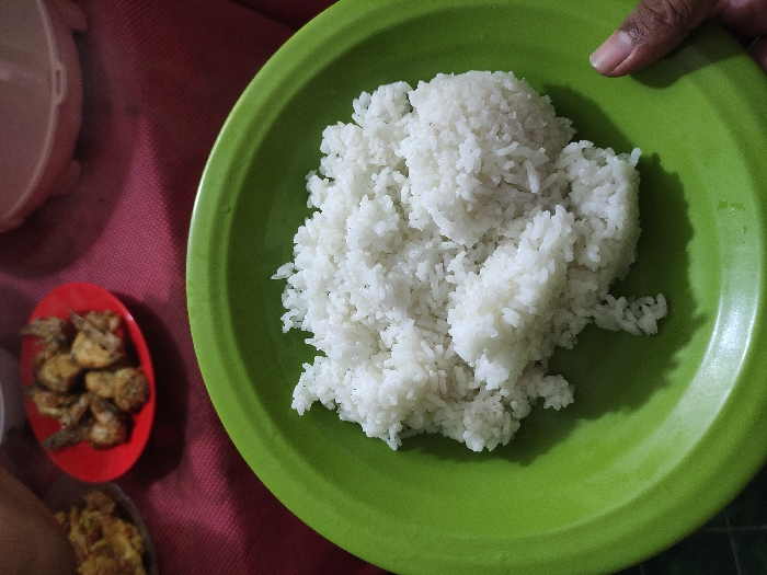 A green plate filled with white rice sits on a red tablecloth. The rice is steaming and appears to have been recently cooked. There is a partial view of a hand to the right of the plate and a blurred background showing two additional plates with food. The coordinates given are for a location in the city of Kecamatan Lawang, Indonesia. The image is likely taken indoors at a kitchen table.