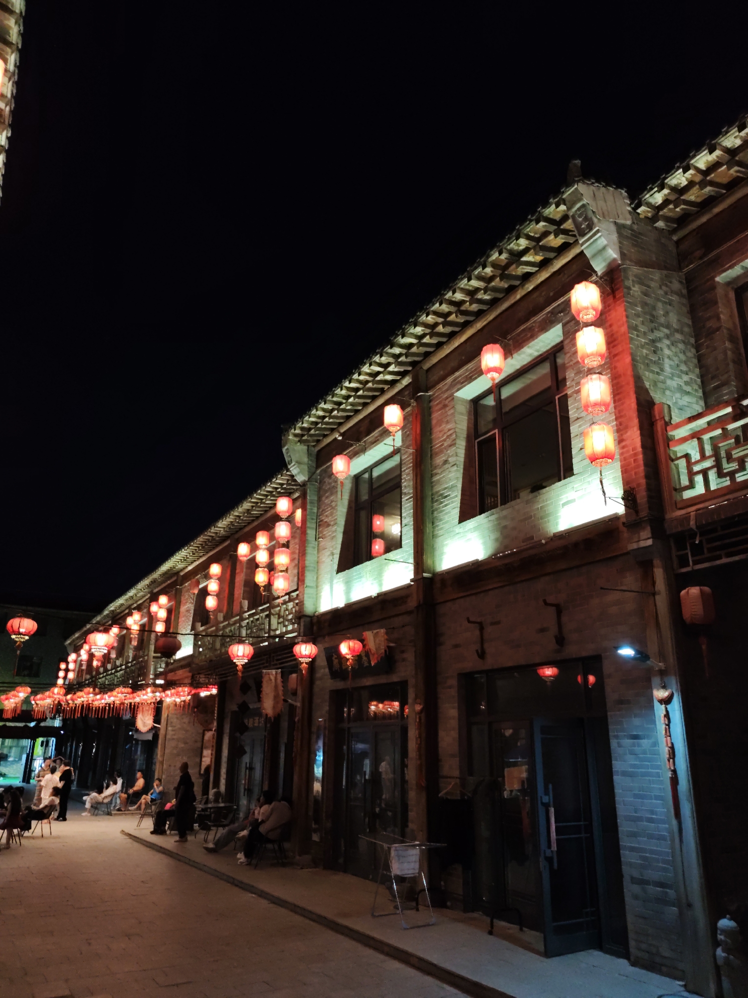 The image depicts a nighttime scene in Lindian, China, showcasing a traditional architectural style along a street lined with red lanterns. The buildings have tiled roofs and brick facades, with decorative elements and illuminated windows. Numerous red lanterns are suspended along the eaves of the buildings, casting a warm glow on the surroundings. Several people are visible in the foreground and midground, some sitting on chairs along the street, while others appear to be standing or walking. The street is paved, and the overall atmosphere suggests a lively evening in a cultural district.
