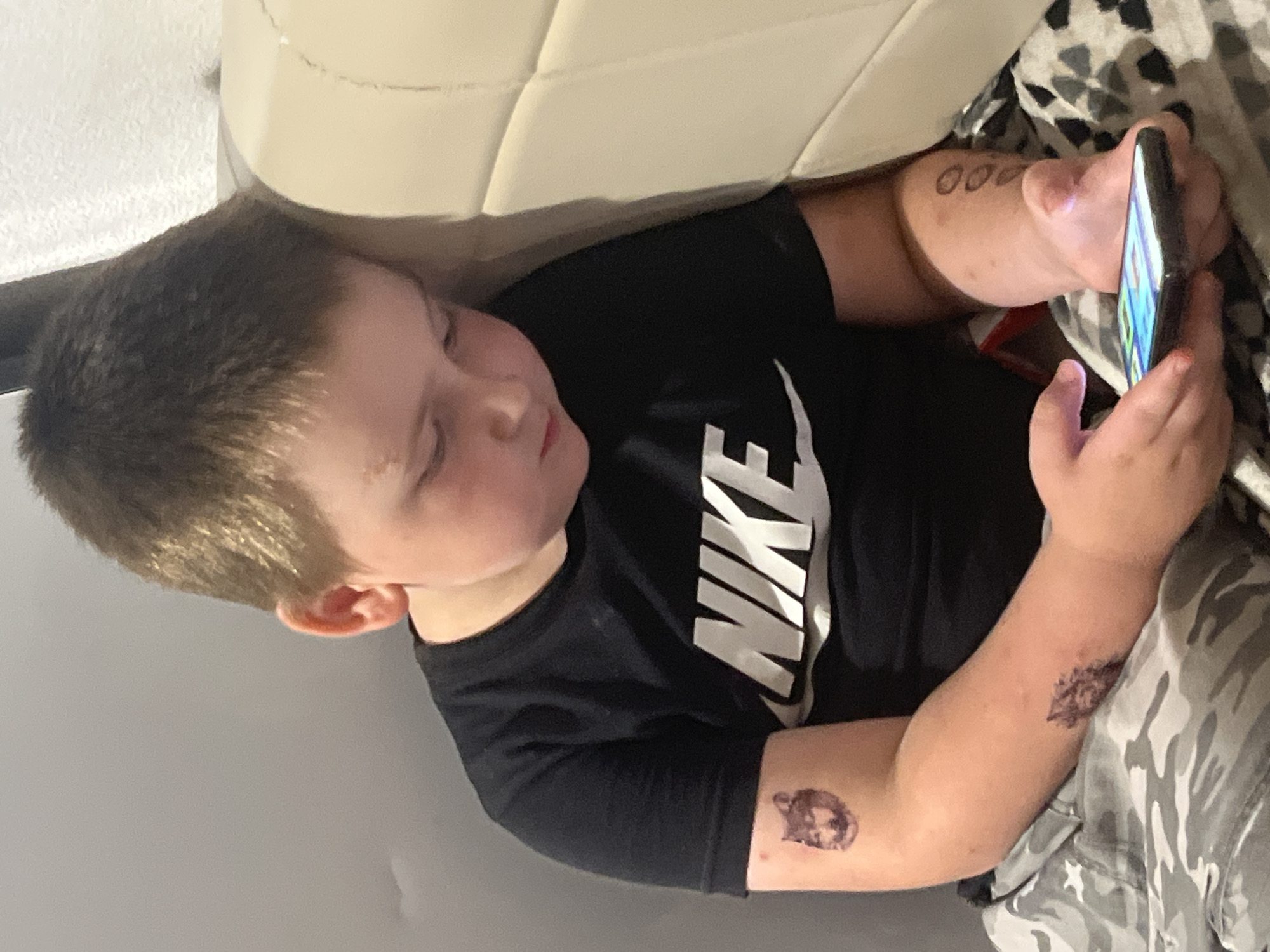 In an indoor setting, likely a bedroom, a young Caucasian boy with short brown hair is leaning against a light-colored headboard, looking down at a smartphone he is holding in his hands. He is wearing a black Nike t-shirt and camouflage patterned pants. The boy has several temporary tattoos on his right arm, including a wolf's head on his upper arm and what appears to be a cluster of circles on his forearm. The image is a medium shot, focusing on the boy and his immediate surroundings. There is no indication of the boy's location within the city of Chelyabinsk, Russia.