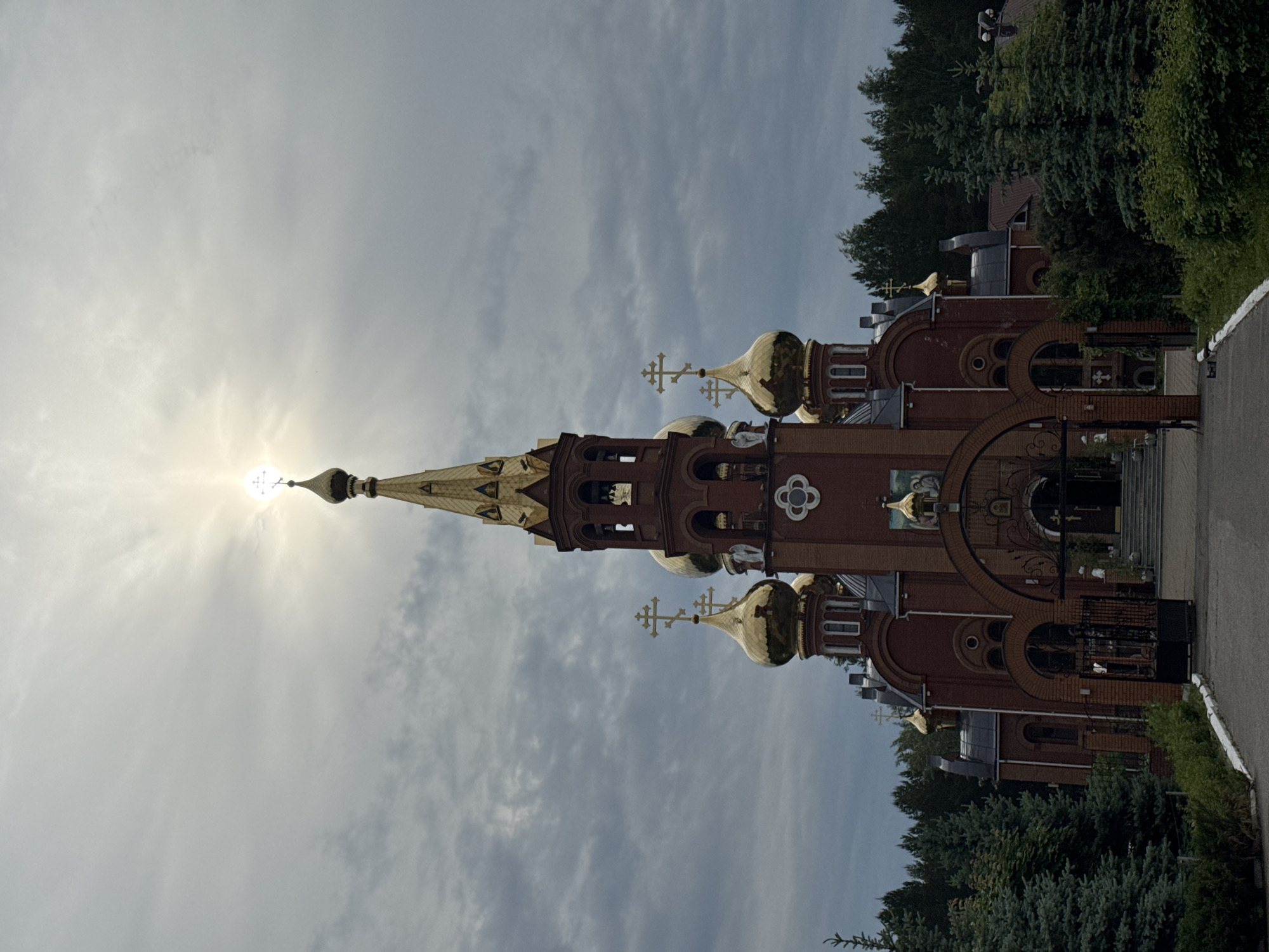 This image, taken in Chaikovsky, Russia, showcases a magnificent Eastern Orthodox church or monastery building, distinguished by its vibrant red brick facade and an array of golden onion domes and spires. Each dome and spire is crowned with a traditional three-barred Orthodox cross.
The tallest spire, prominently positioned towards the upper left, features a cross directly backlit by the sun, creating a brilliant sunburst effect and a strong lens flare that illuminates the sky. The building exhibits intricate architectural details, including gracefully arched windows and doorways, ornamental brick patterns, and what appears to be an outdoor icon or mural integrated into its upper facade. Ornate dark metalwork, possibly a gate or fencing, is visible near the building's entrance.
A paved pathway or road runs along the right side of the structure, edged by neatly trimmed low hedges. Dense green trees and various bushes frame the right edge of the image and extend behind the building, indicating a landscaped setting. The sky above is partly cloudy, with the bright sun breaking through.
The scene is tranquil and static, highlighting the grandeur of the religious architecture under bright, diffused daylight. No individuals or specific events are depicted. The architectural style clearly identifies it as an Orthodox place of worship, typical of the region.