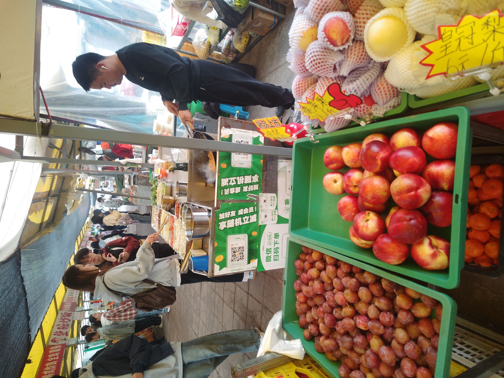 A street food market scene in Shapingba District, China, features a vendor wearing black clothing and an earpiece preparing food at a stall. Several customers are browsing at the stall. There are containers filled with different fruits, including peaches, plums, and Asian pears. A promotional sign in Chinese advertises a discount for adding the vendor as a friend on WeChat and scanning a QR code. Prices are displayed on yellow signs. The area appears to be crowded with shoppers.