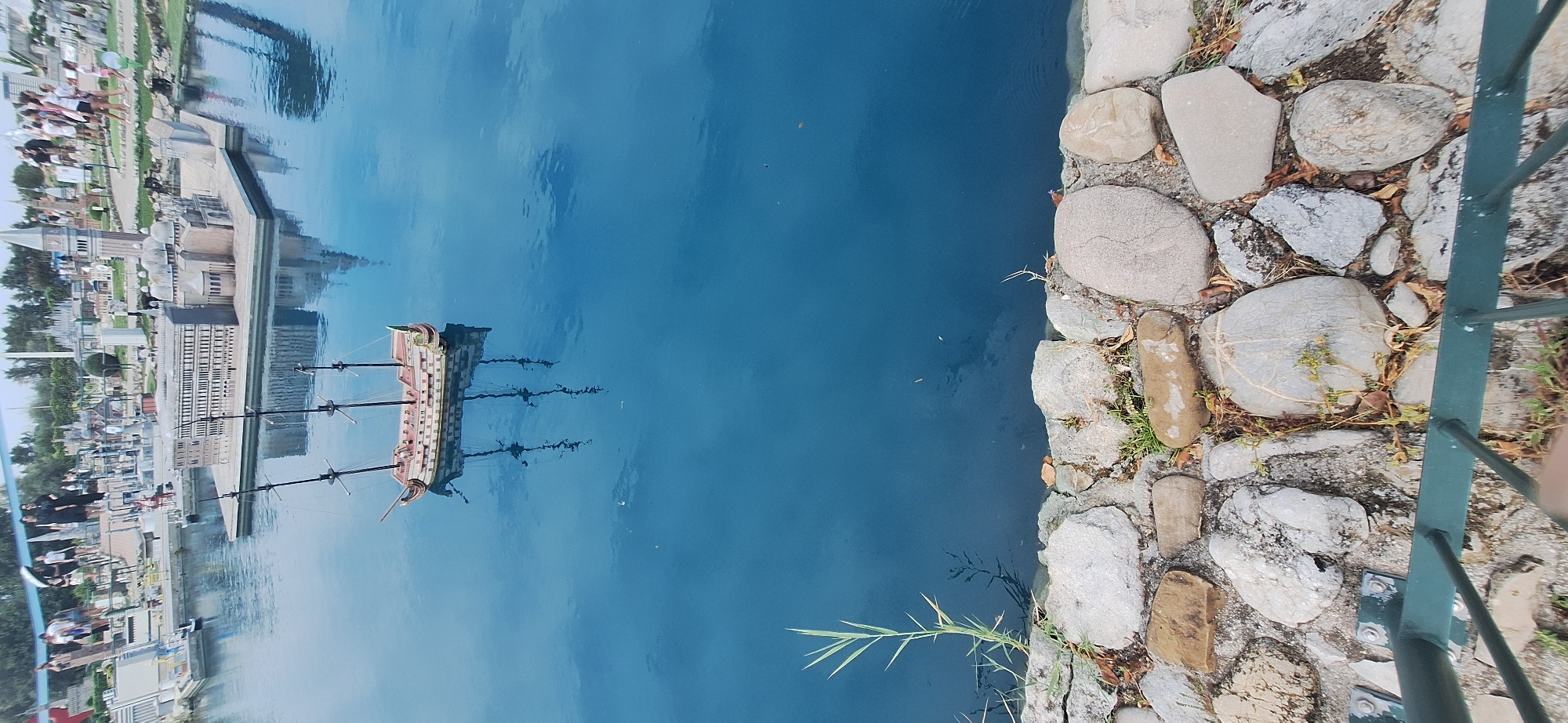 The image shows a miniature model of Venice, Italy, located in the Italia in Miniatura park in Rimini. A model of a tall sailing ship is floating in the water in the foreground. In the background, miniature buildings representing Venice's landmarks, such as St. Mark's Basilica and the Doge's Palace, are visible. Several miniature figures of people are also present, walking around the model city. The water is a deep blue, and its surface reflects the sky and the surrounding models. The foreground of the image shows a stone embankment with some grass and dry leaves. A green metal railing is visible at the very bottom of the image, indicating a viewing area.