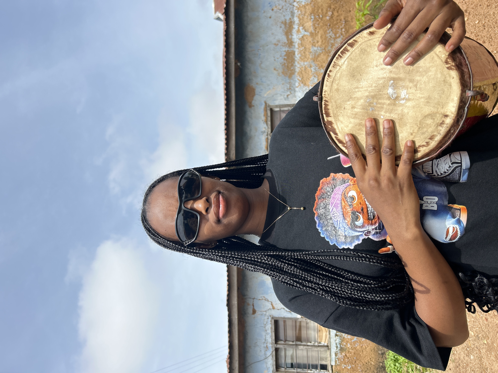 A young woman with long black braids and black sunglasses stands outdoors, holding a small drum. She is wearing a black t-shirt with a colorful graphic of a cartoon character. Behind her is a distressed, light blue building with a weathered facade and visible cracks. The sky above is blue with scattered white clouds. The ground appears to be dirt or gravel. The scene is likely within a residential area in Jos, Nigeria, suggested by the architectural style of the building and the outdoor setting typical of the region.