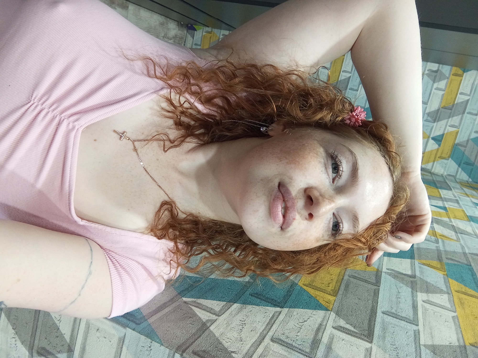 A young woman with red curly hair lies on a surface with a geometric pattern of gray, blue, and yellow. She is wearing a light pink top and a delicate gold necklace with a cross pendant. A small pink flower is tucked into her hair near her ear. She has fair skin with freckles and is looking towards the camera. A faint circular tattoo is visible on her left arm. The image appears to be taken indoors.
The location in Yurgamysh, Russia, cannot be determined from the image.
