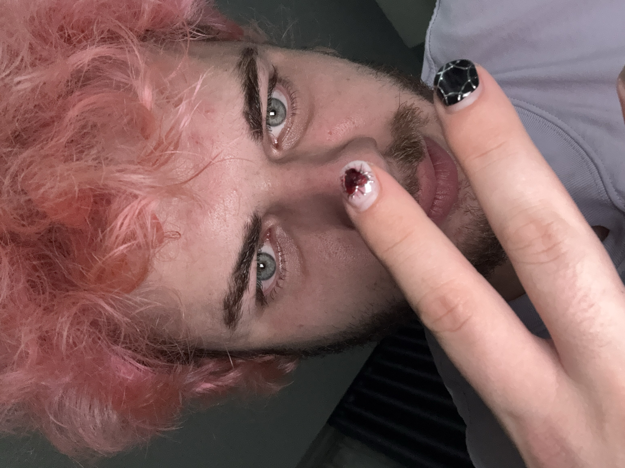 A man with pink hair is positioned upside down in the frame. He has light skin, grey eyes, and a short beard. One hand is raised, with the middle finger pointed towards his nose. His nails are decorated; the nail on the middle finger features a design with red and silver accents, while the nail on the ring finger has a black and silver geometric pattern. He is wearing a light-colored shirt. The background is indistinct and appears to be a room with a wall and possibly a slatted surface. The image provides no information about Đà Nẵng, Vietnam.