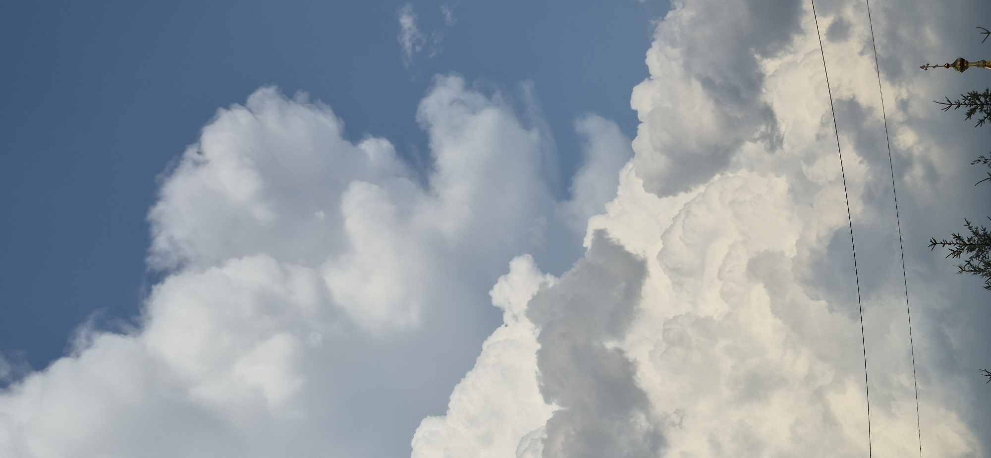 The media file is a low-angle shot of a dramatic sky filled with large, billowing cumulus clouds against a bright blue sky. The clouds are illuminated by sunlight, with some areas appearing bright white and others showing shades of gray, suggesting depth and volume. The composition is dominated by the clouds, which fill most of the frame, creating a sense of awe and grandeur.
In the upper right portion of the image, there are thin, dark wires running vertically, characteristic of power or utility lines. Also in the upper right, a portion of a decorative architectural element, possibly a spire or finial with intricate detailing and some gold coloring, is visible, along with dark green foliage from a tree. These elements suggest a human-made landscape intersecting with the natural sky.
The overall impression is of a bright, possibly late morning or afternoon sky, with dynamic cloud formations. The scene is serene yet powerful, with the interplay of light and shadow on the clouds adding to the visual drama. The presence of utility lines and architectural detail grounds the sky in a specific, potentially urban or semi-urban, environment. The location context of Ussurijsk, Russia, adds a geographical marker to the scene. There is no visible text within the image.