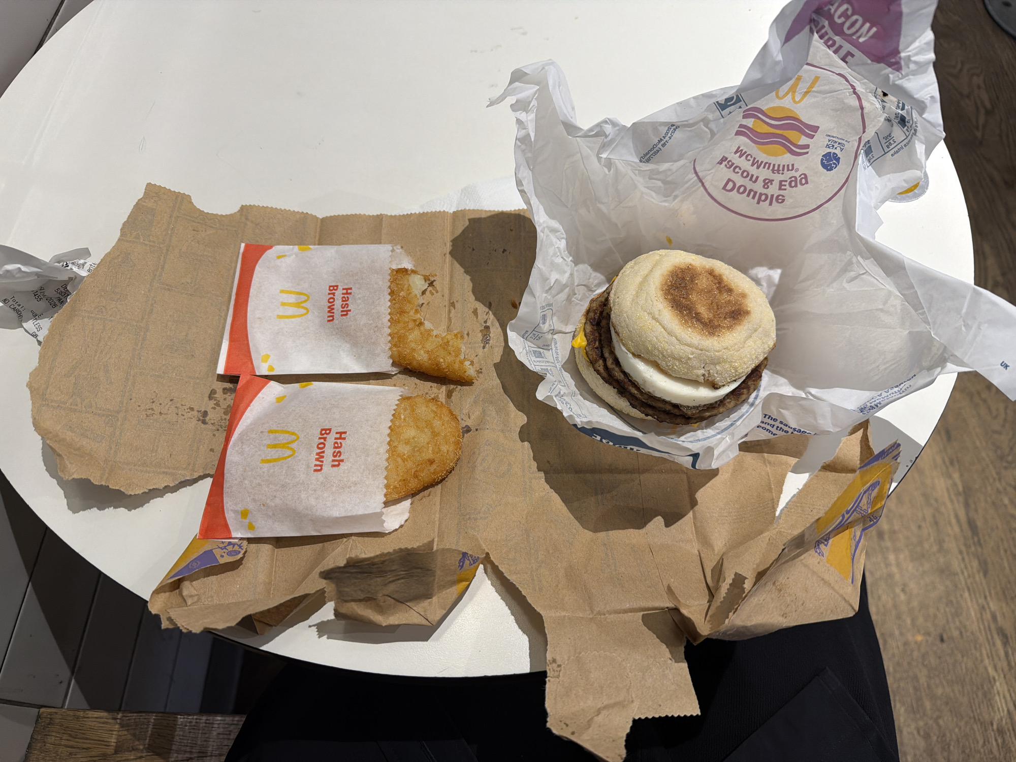 An overhead shot captures a partially consumed McDonald's breakfast meal on a white, round table. The scene is located within a McDonald's establishment in London, United Kingdom, identifiable by the branded packaging, including a "McMuffin Bacon & Egg Donople" wrapper and hash brown sleeves. Two hash browns are present; one remains intact in its white paper sleeve, while the second has a visible bite taken from its top-right portion and is also in its branded sleeve. A partially unwrapped McMuffin, containing an egg and a sausage patty, sits to the lower right. All food items rest upon a crumpled, grease-stained brown paper McDonald's bag. The immediate environment includes a dark, possibly fabric-covered surface on the left, next to a vertical wooden panel, suggesting a nearby seat or wall element. Bright lighting from above casts distinct shadows from the food and packaging onto the table. No individuals are visible.